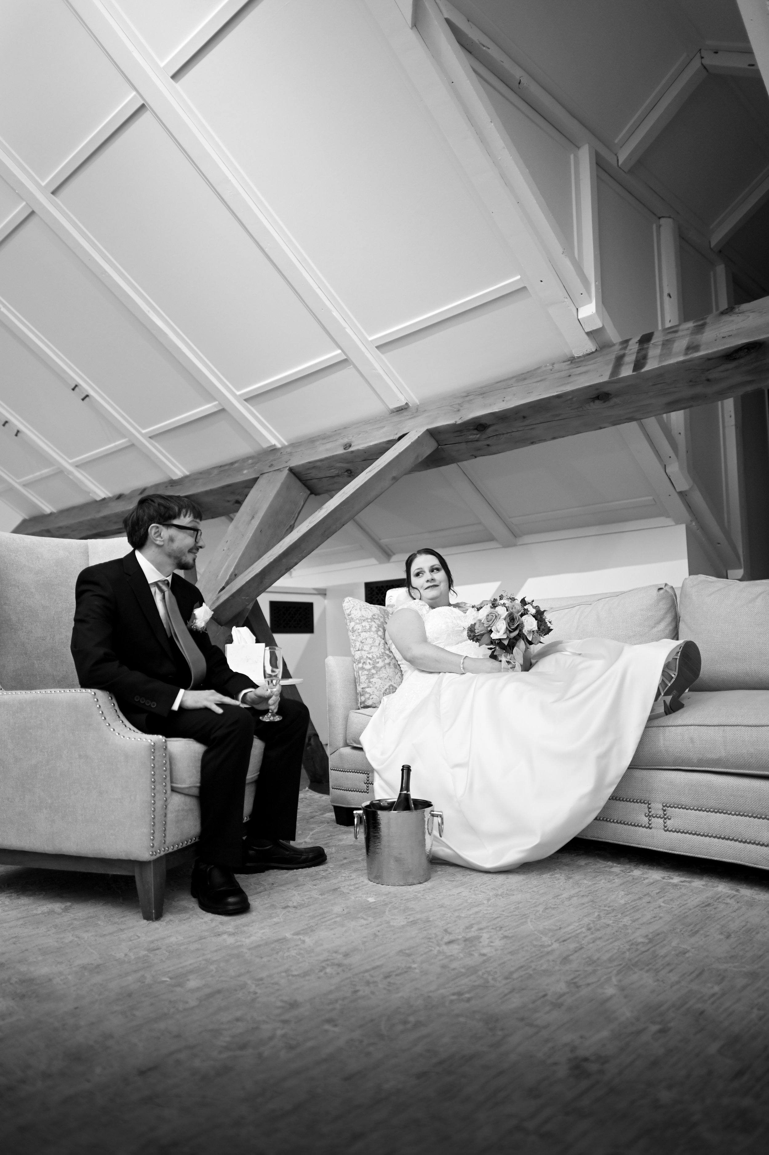 A black and white photo of a bride in a wedding dress sitting on a sofa, holding a bouquet, with a groom in a suit sitting on a separate couch in front of her, holding a glass of champagne, in a room with sloped ceiling and exposed wooden beams.