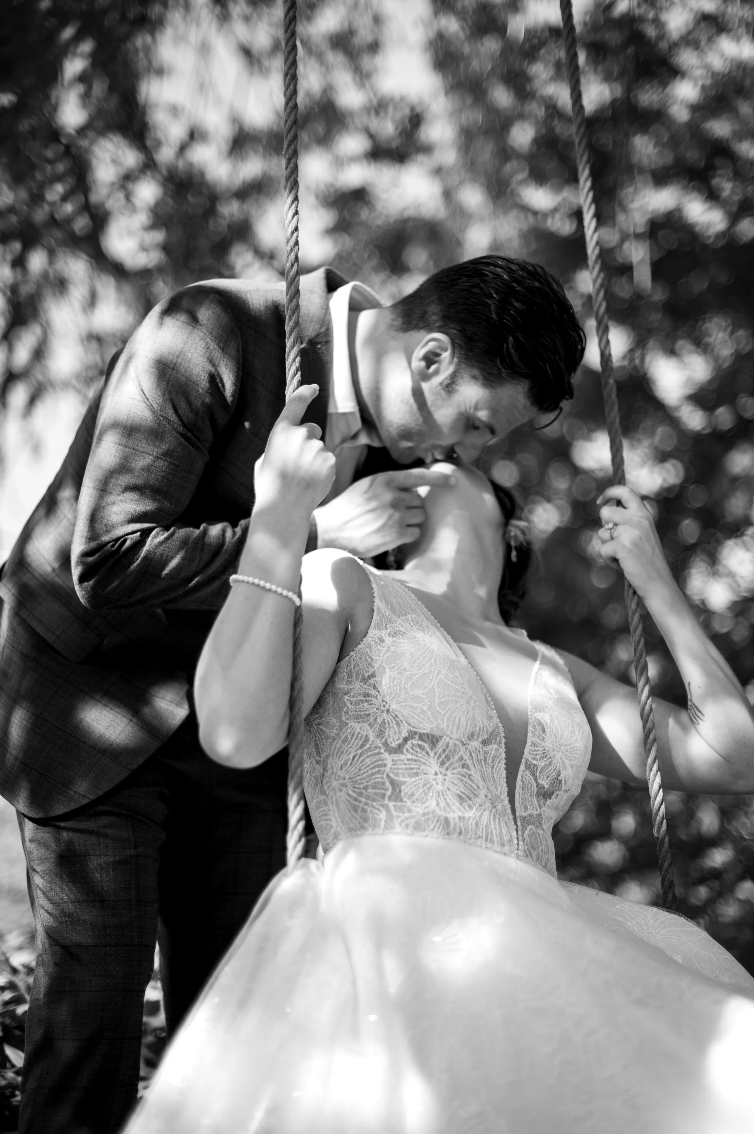 A black-and-white photo of a man and woman on a swing. The man, dressed in a plaid suit, is leaning in to kiss or whisper to the woman, who is wearing a lace dress. The swing's ropes are visible, and the background consists of blurred trees.