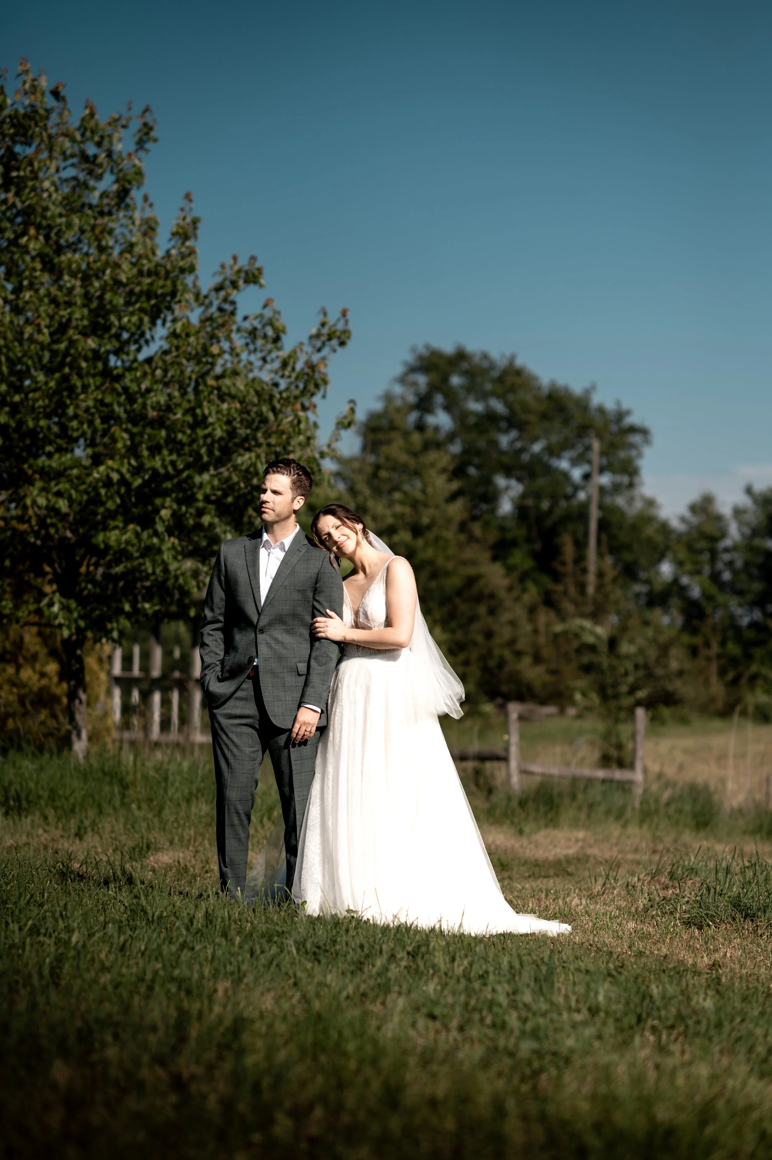 A bride and groom standing together outdoors in a grassy area with trees and a wooden fence in the background, under a clear blue sky.