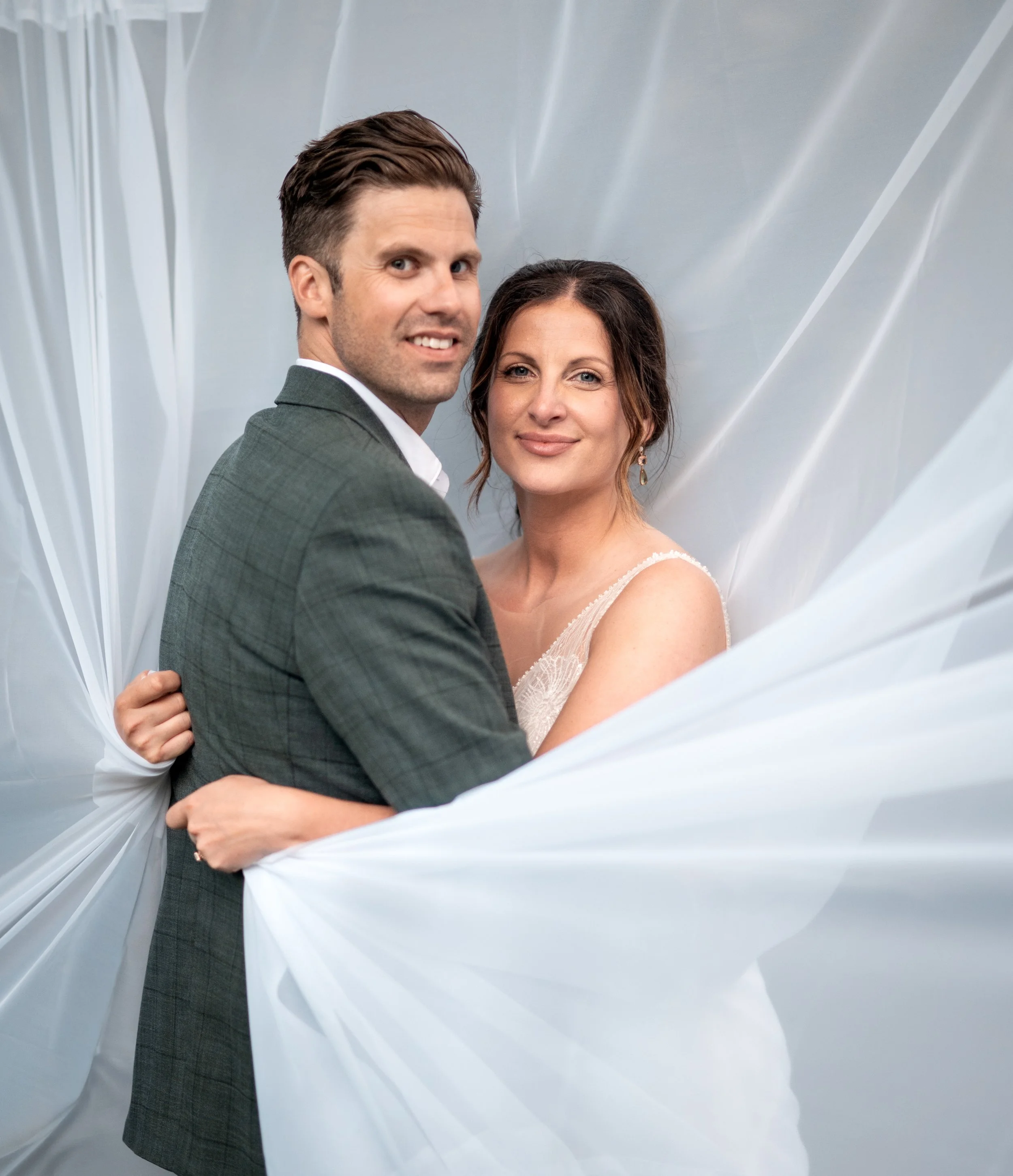 A smiling couple dressed in wedding attire, the man in a suit and the woman in a white dress, posing together in front of a soft, draped white background.