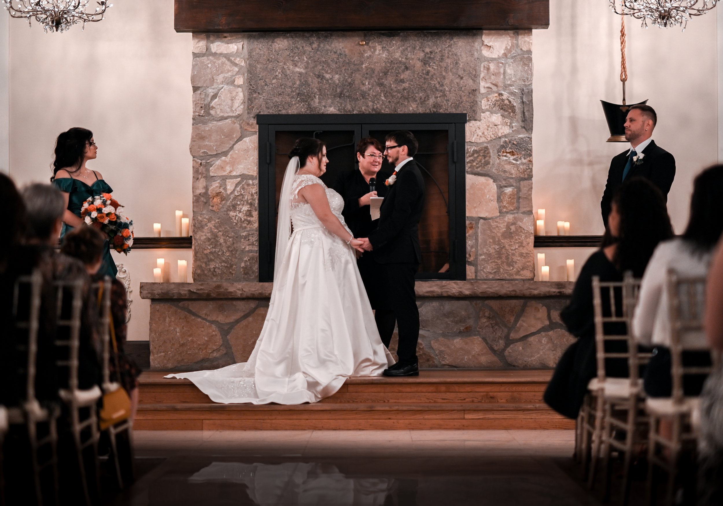 A wedding ceremony taking place in front of a stone fireplace, with the bride and groom holding hands and exchanging vows. The officiant stands between them, and a bridesmaid is holding a bouquet on the left. Guests seated on either side are watching