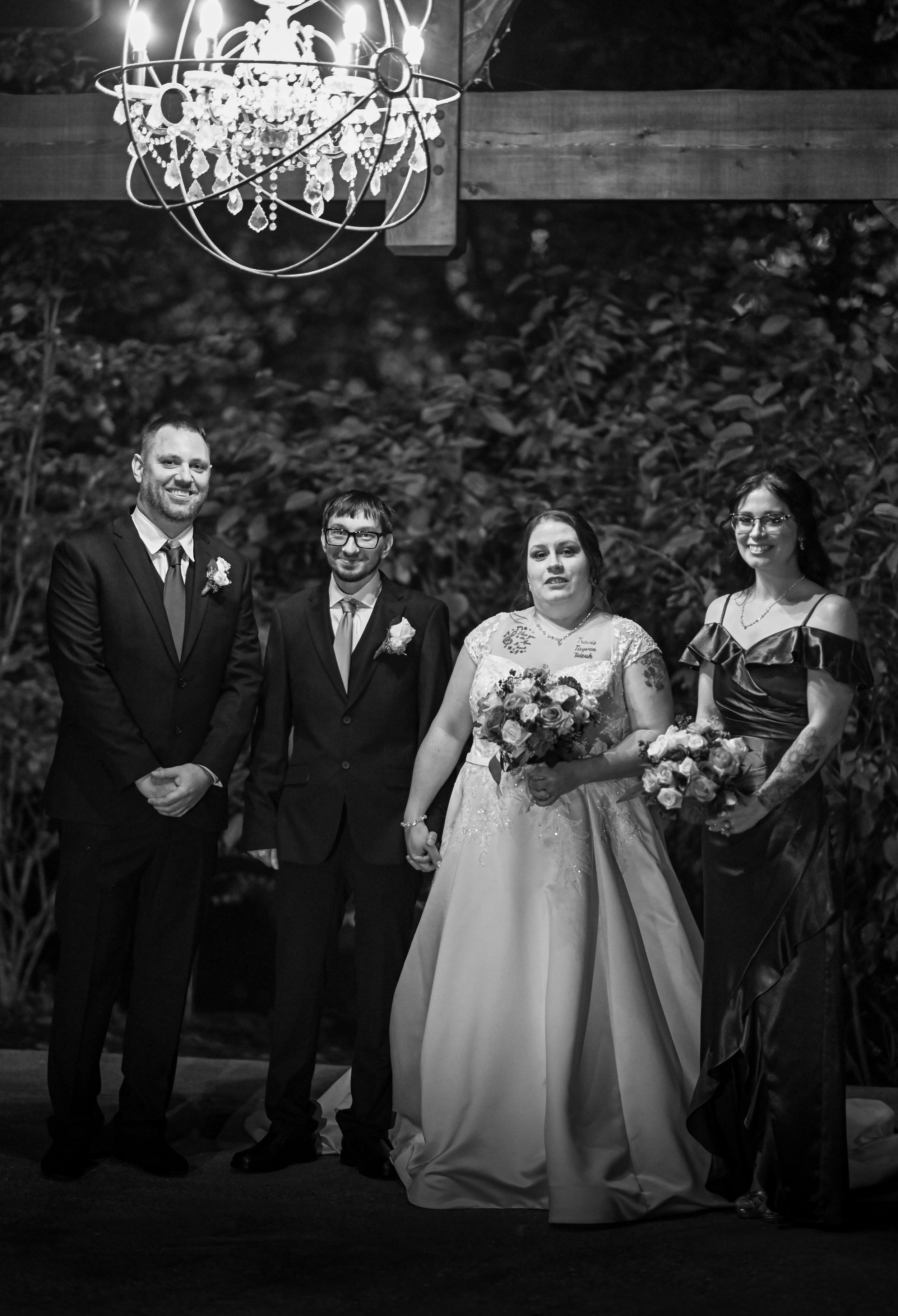 Black and white photo of four people at a wedding ceremony under a chandelier, with two men in suits and two women in dresses, standing outdoors against a backdrop of trees and foliage.
