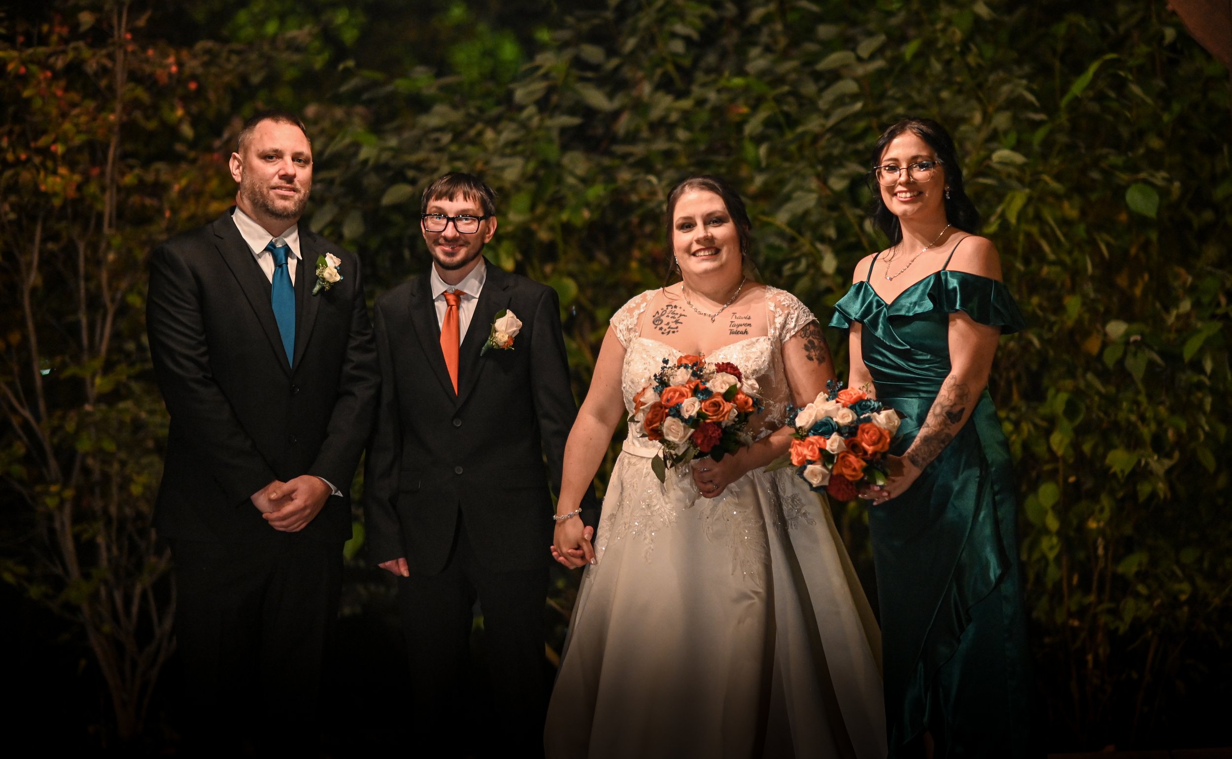 Group of five people at a wedding, standing outdoors at night in front of green foliage. The bride, in a white gown and holding a bouquet, is in the center. To her left are two men in suits with boutonnières, and to her right are two women, one in a 