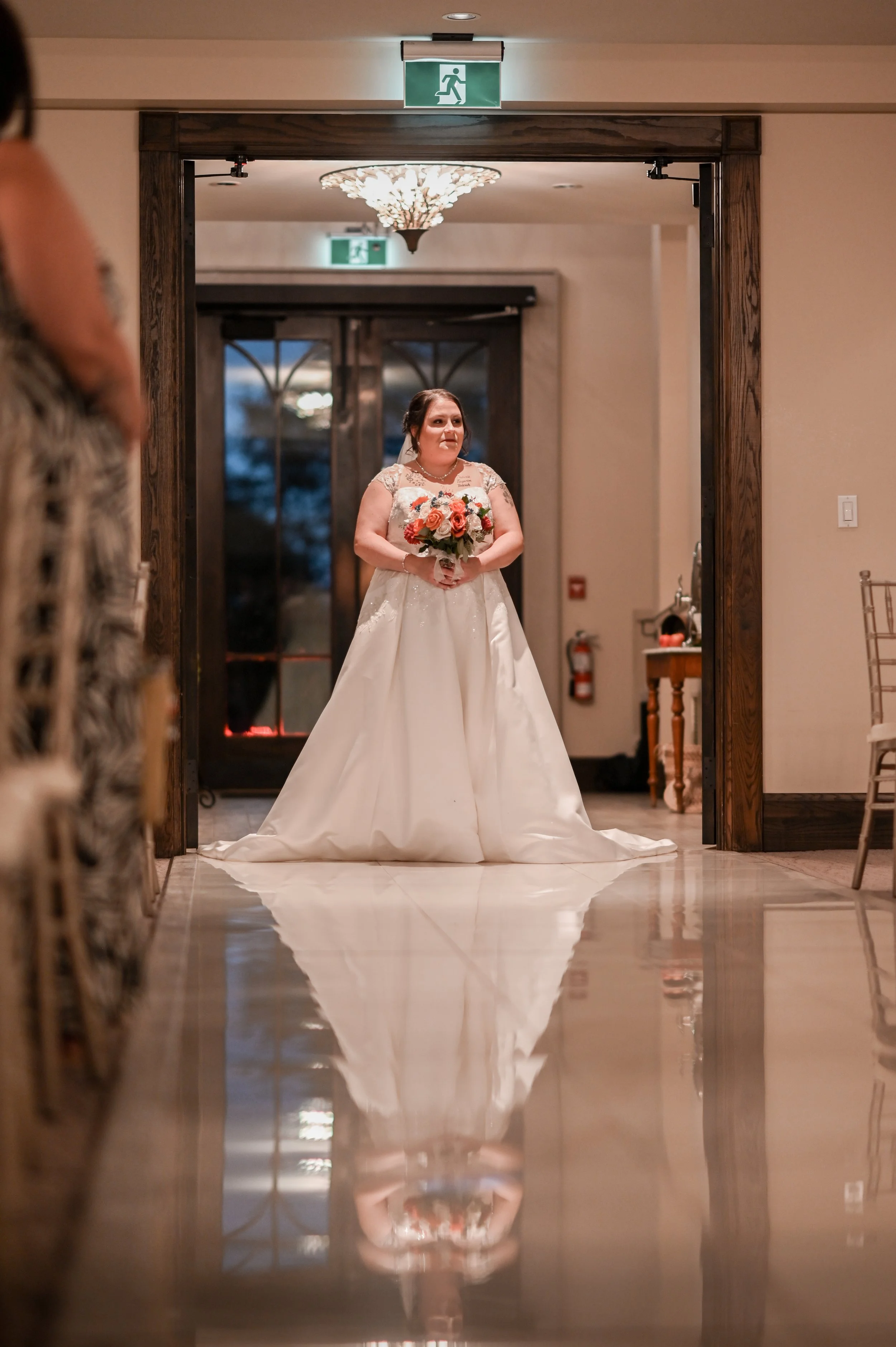 Bride in a white wedding gown holding a bouquet, standing in a doorway with her reflection visible on the shiny floor, indoors at a wedding reception.