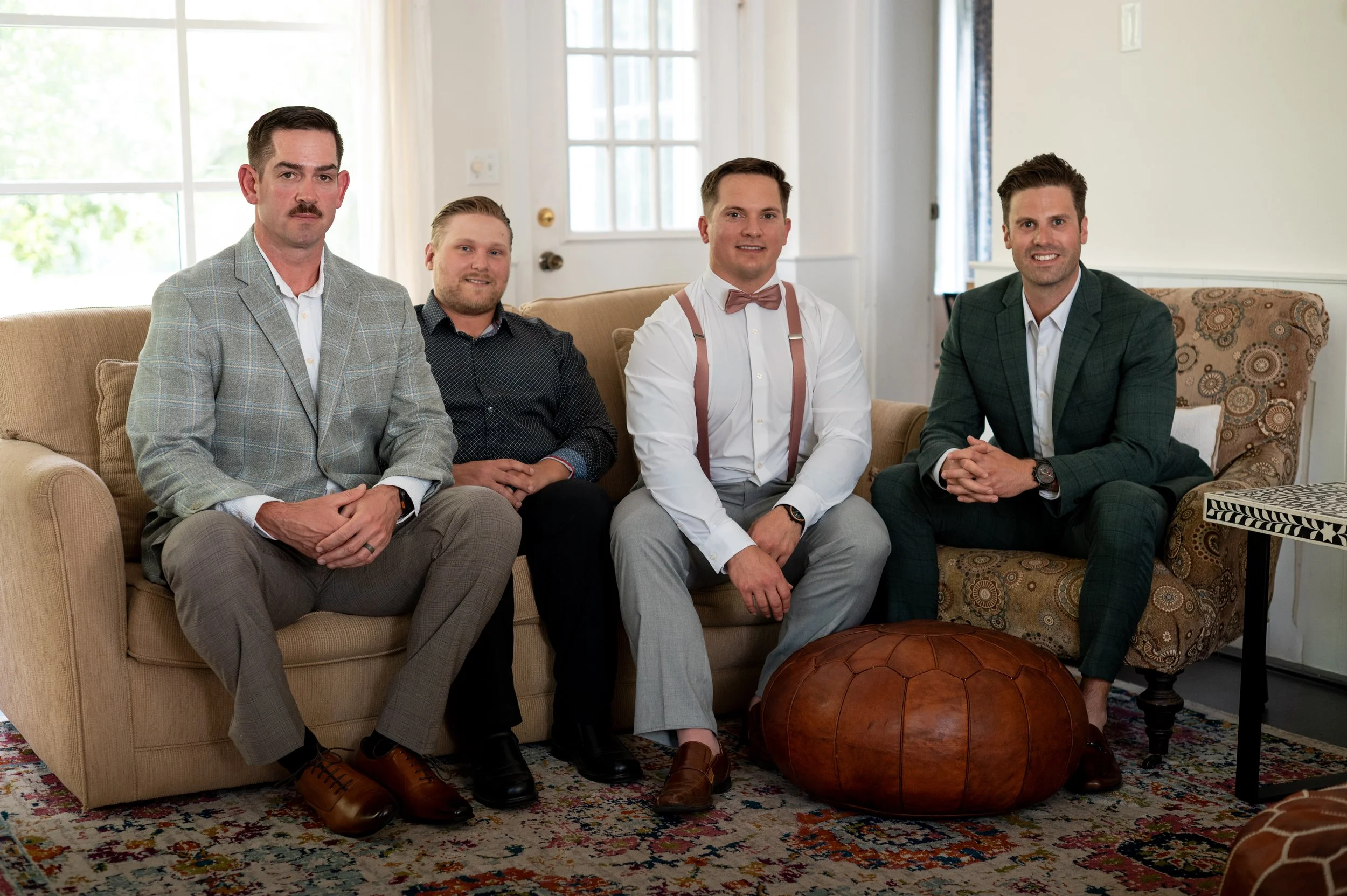 Four men sitting on a beige couch in a well-lit room with large windows, dressed in stylish suits and shirts, posing for a photo with relaxed expressions.
