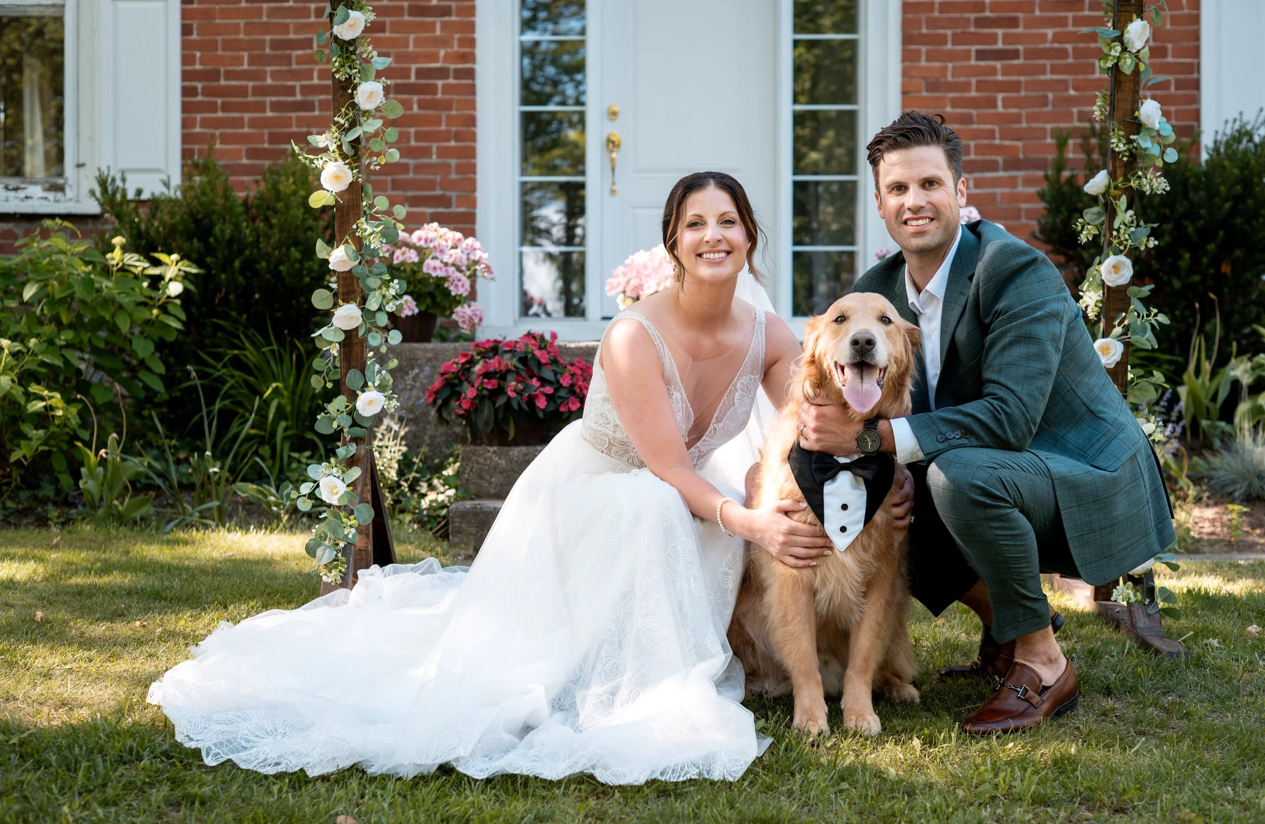 A smiling bride and groom with a large Golden Retriever dressed in a tuxedo, posing for a photo outside a brick house with a white door, surrounded by flowers and greenery, during a wedding celebration.