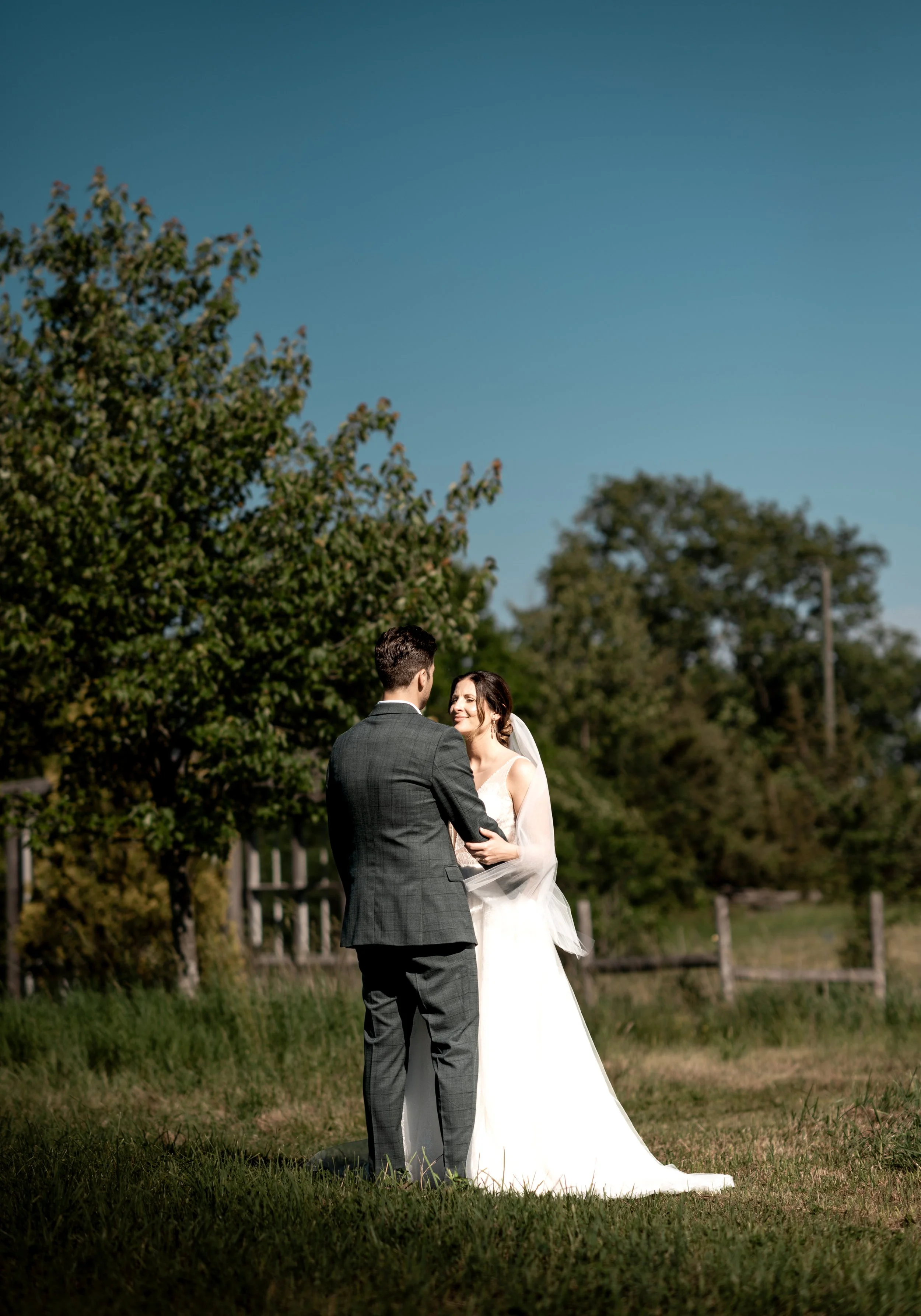 A couple dressed in wedding attire standing close to each other outdoors, with trees and a fence in the background under a clear blue sky.