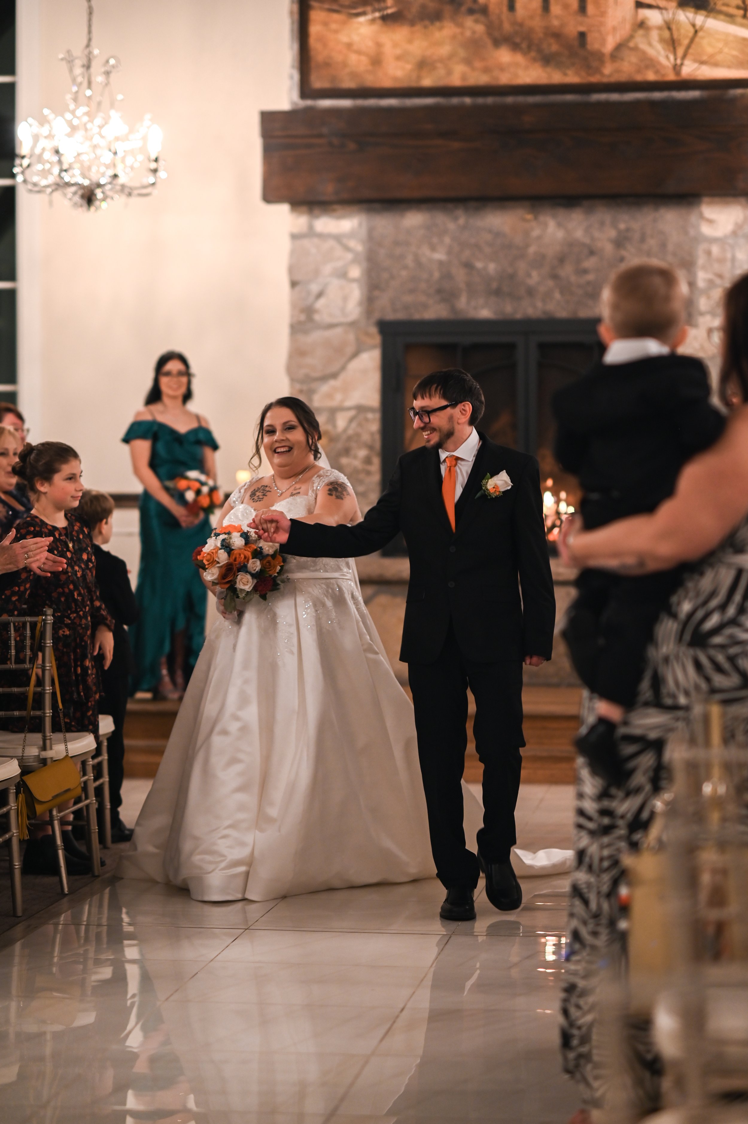 A bride and groom walk down the aisle at their wedding reception, smiling and arm in arm, with guests applauding on either side in a decorated hall with a large stone fireplace and artwork above.