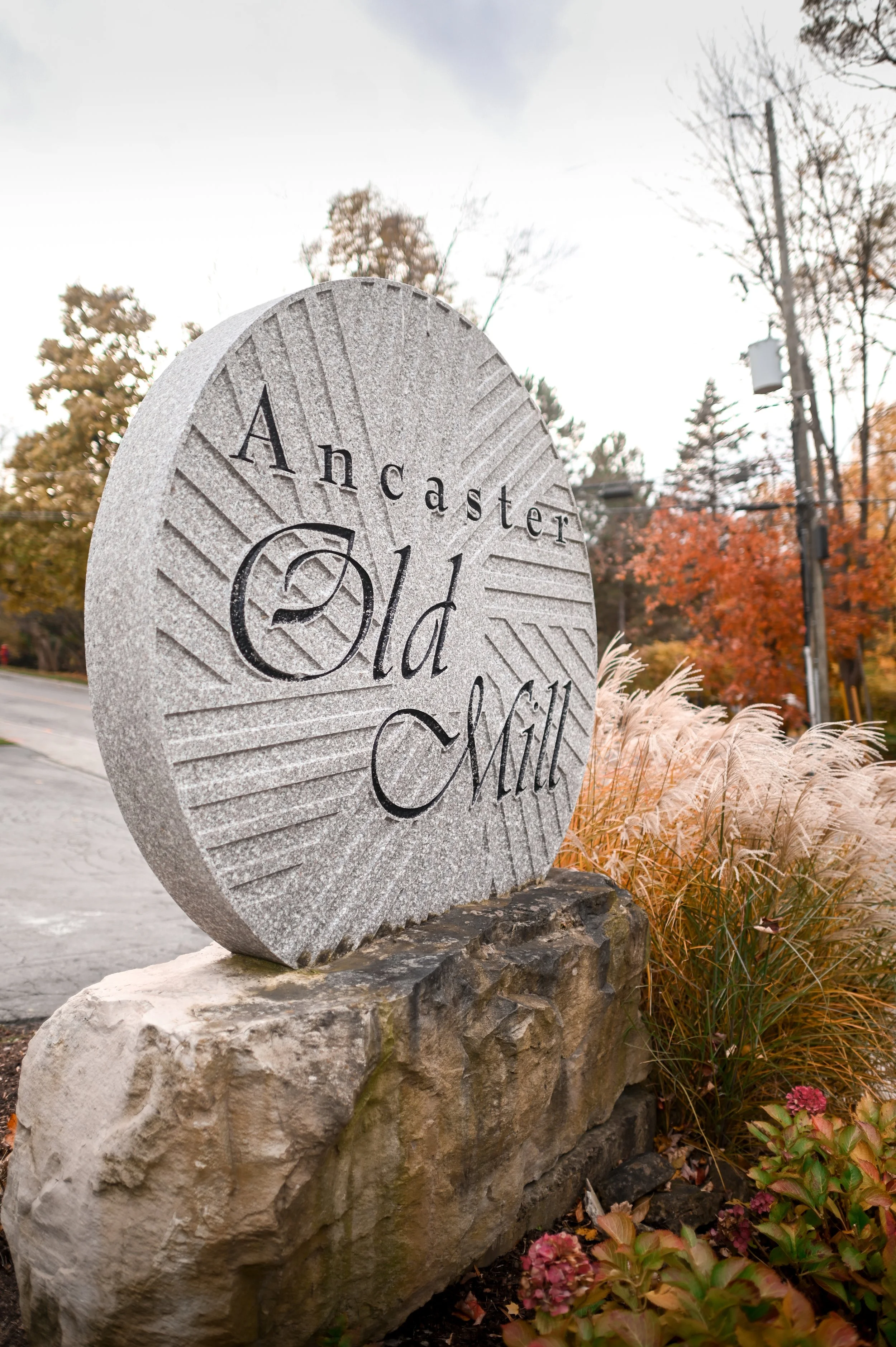 Stone sign with the text "Lancaster Old Mill", surrounded by autumn-colored trees and ornamental grasses.