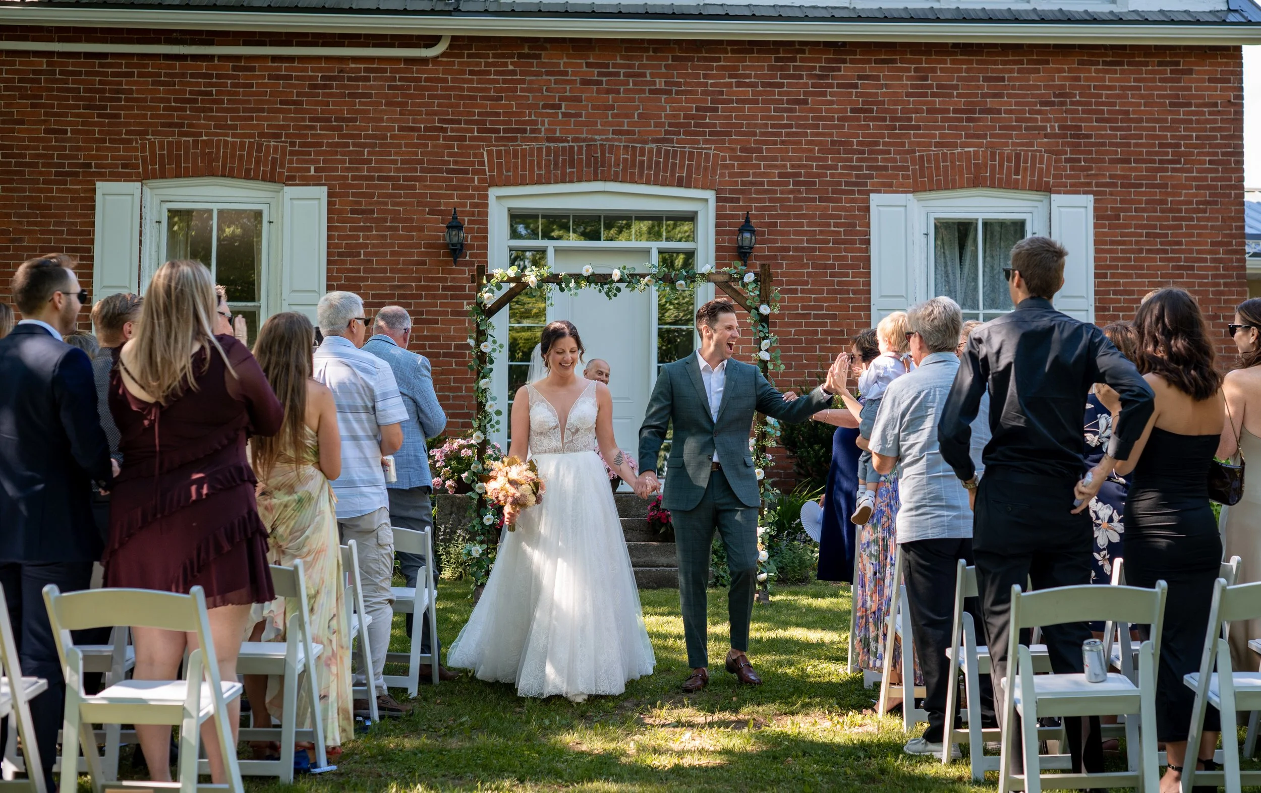 A couple getting married outdoors in front of a brick house, surrounded by friends and family during their wedding ceremony.