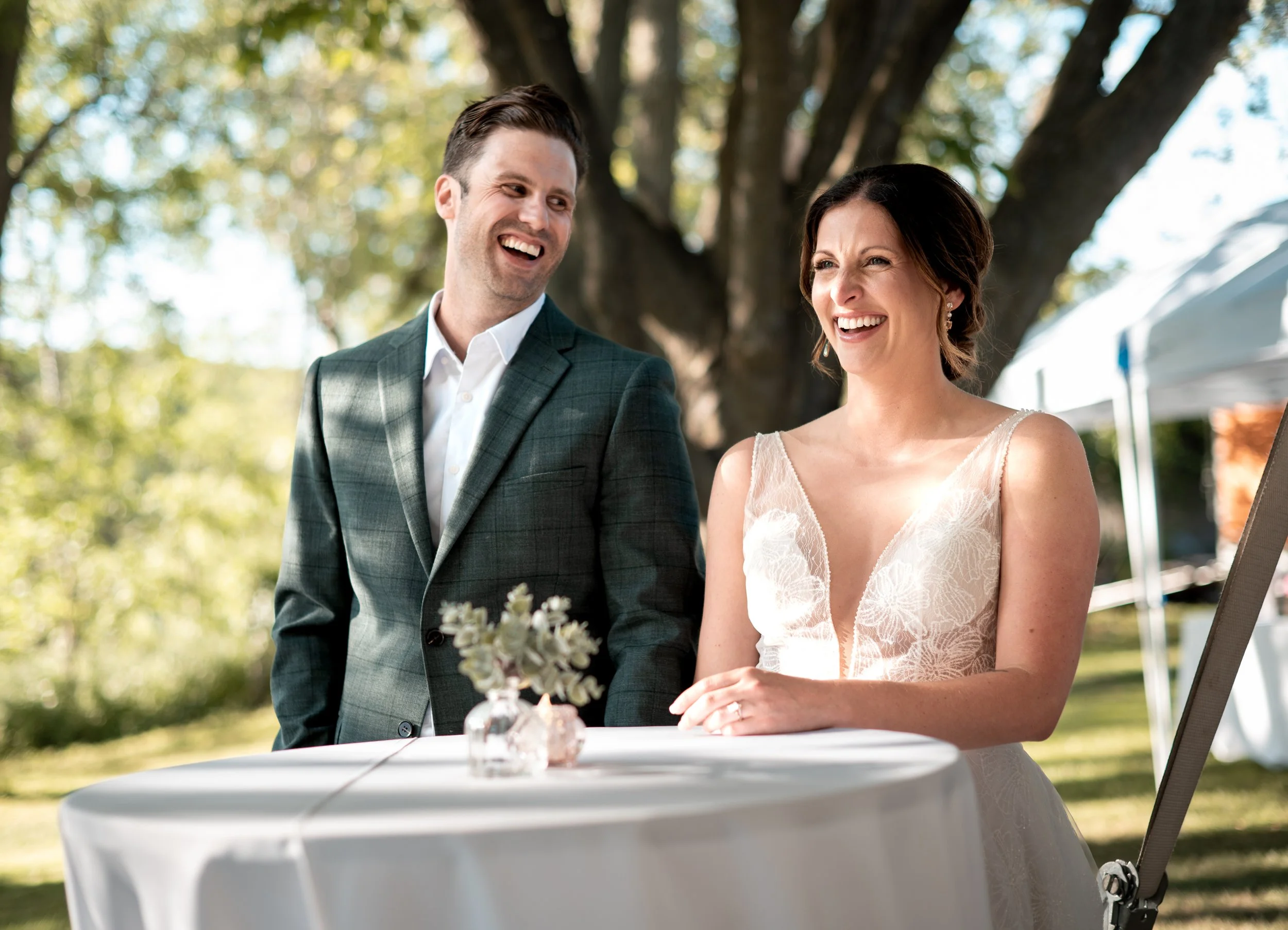A smiling man and woman, dressed in wedding attire, standing outside under a large tree during daytime, with a table covered with a white cloth in front of them and a small floral arrangement on it.