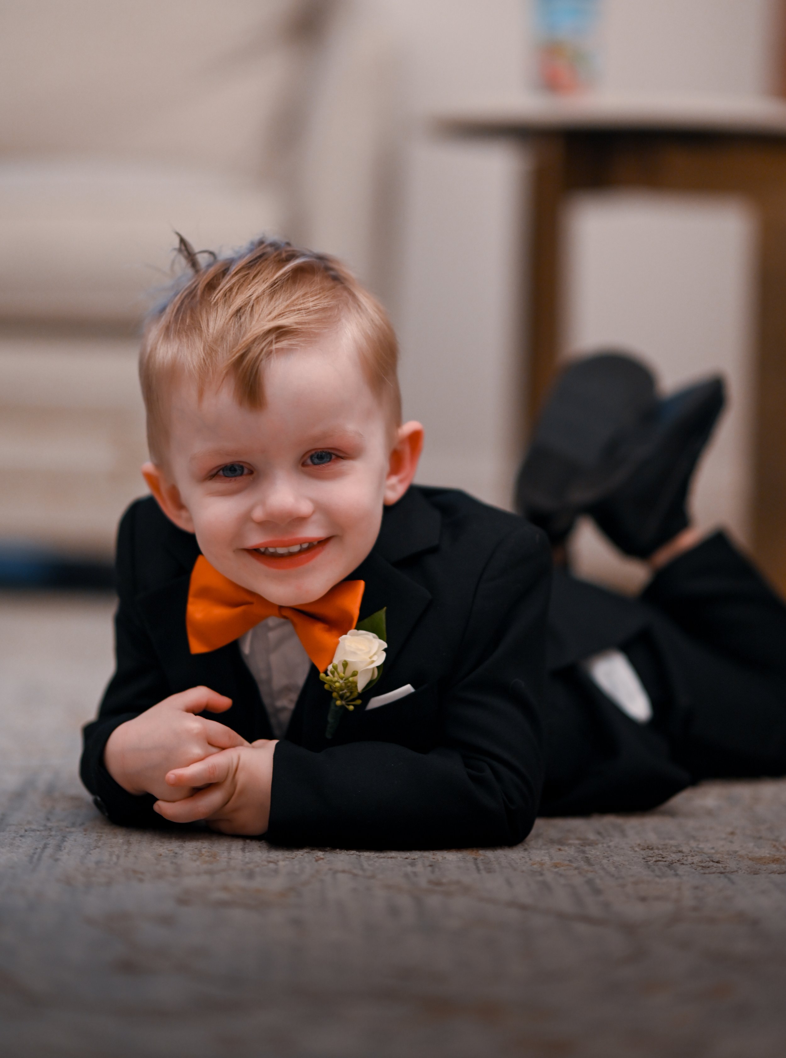 A young boy with red hair and blue eyes is lying on a carpeted floor. He is dressed in a black suit with an orange bow tie, white shirt, and a boutonnière. He is smiling and looking at the camera. In the background, there is a beige chair and a small