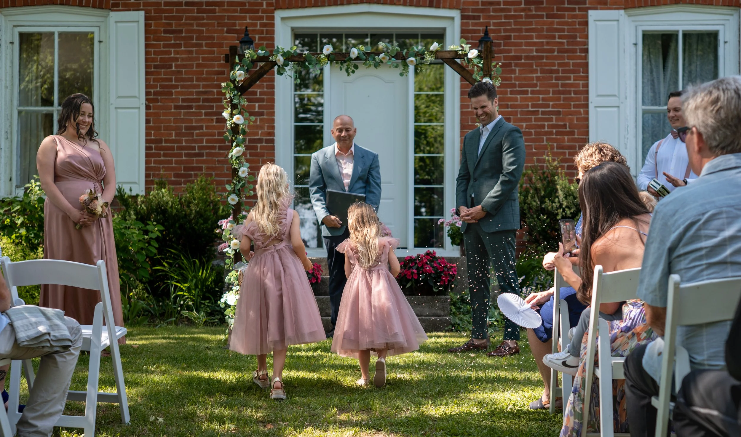 A wedding ceremony outdoors in front of a brick house with white-framed windows. Three flower girls in pink dresses walk down the aisle, and the officiant and groom smile as they watch them. Guests sit on white chairs, some taking photos or videos, w
