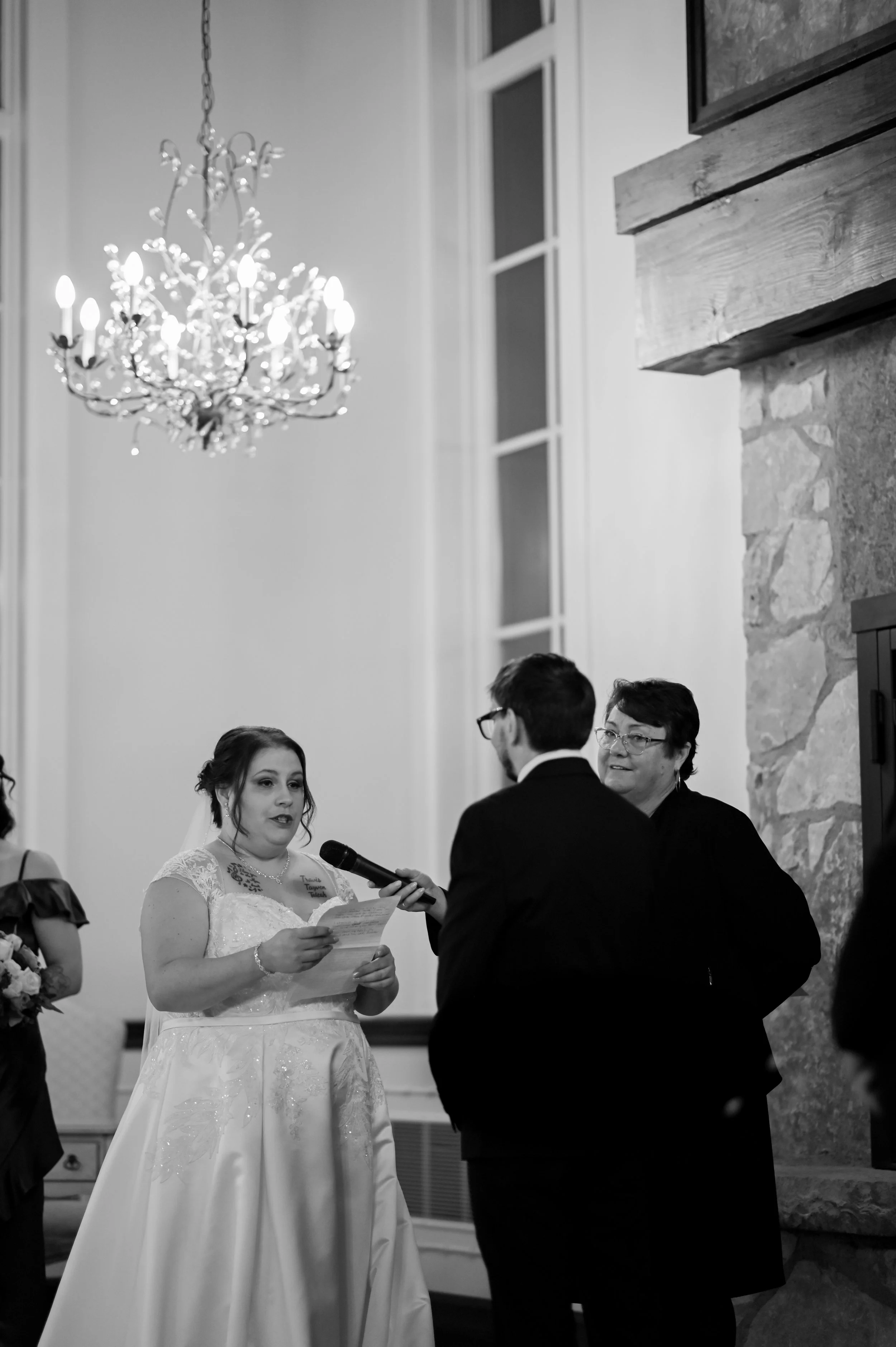 A black and white photo of a bride reading vows into a microphone during a wedding ceremony, with an officiant and a man standing together.