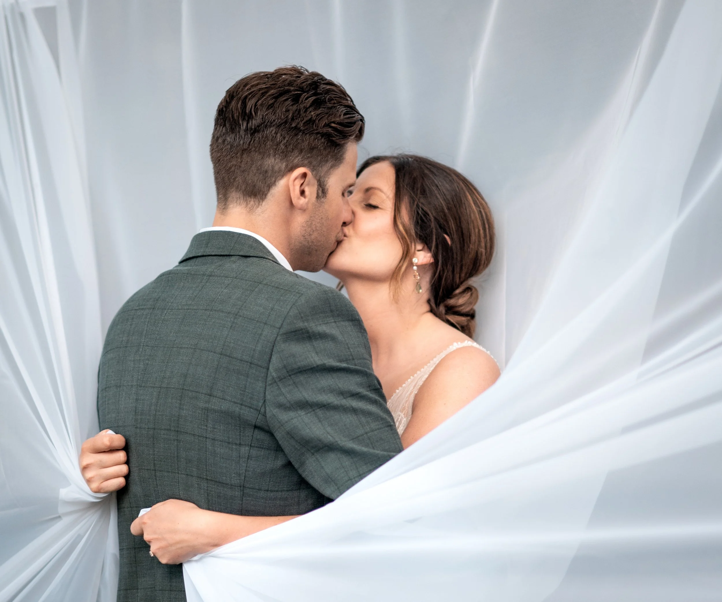 A man and woman sharing a kiss in a close embrace, framed by a white fabric or curtain-like background.