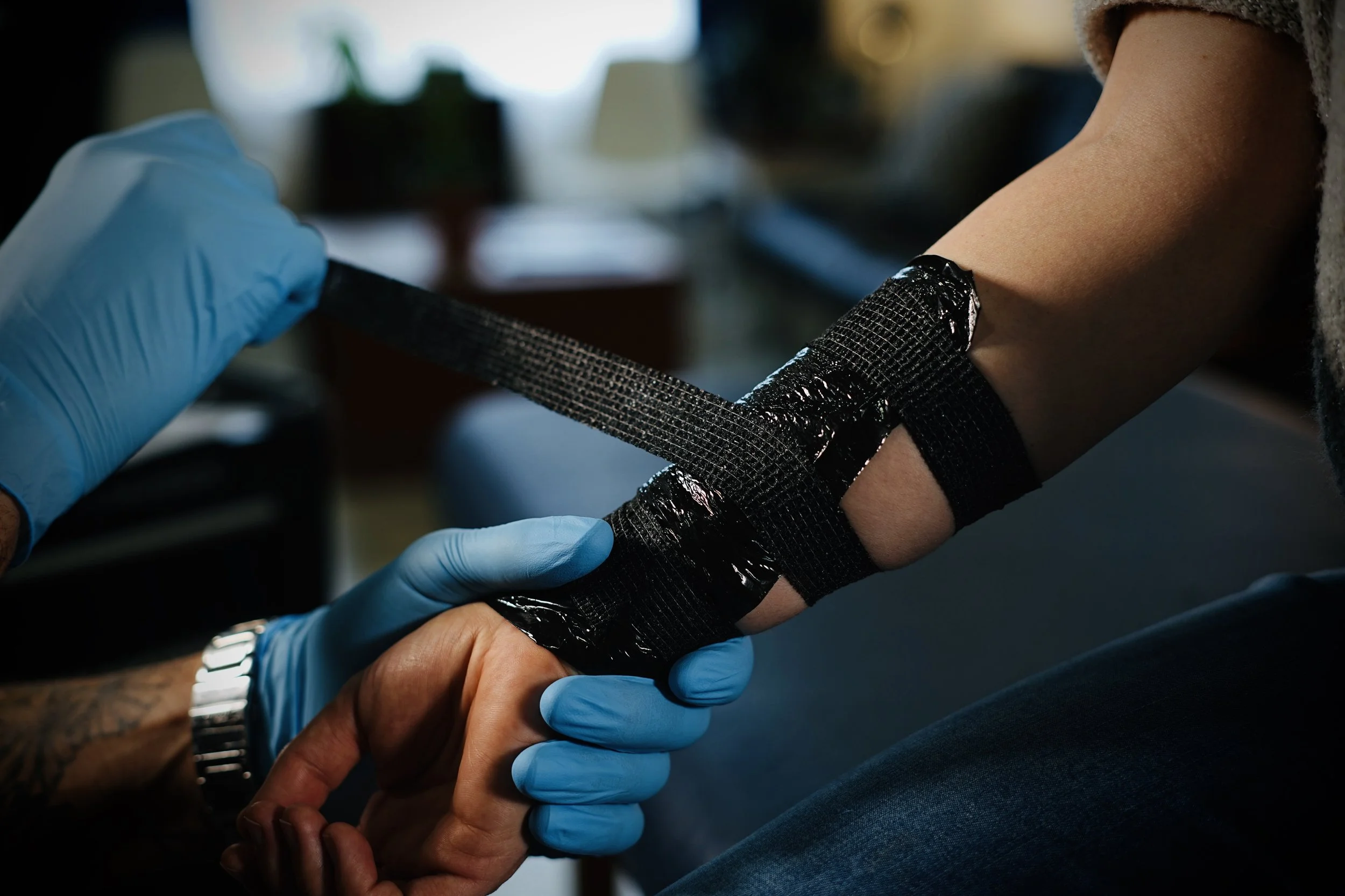 A person receiving a tattoo on their forearm while a tattoo artist, wearing blue gloves, prepares and wraps the fresh tattoo with black adhesive tape.