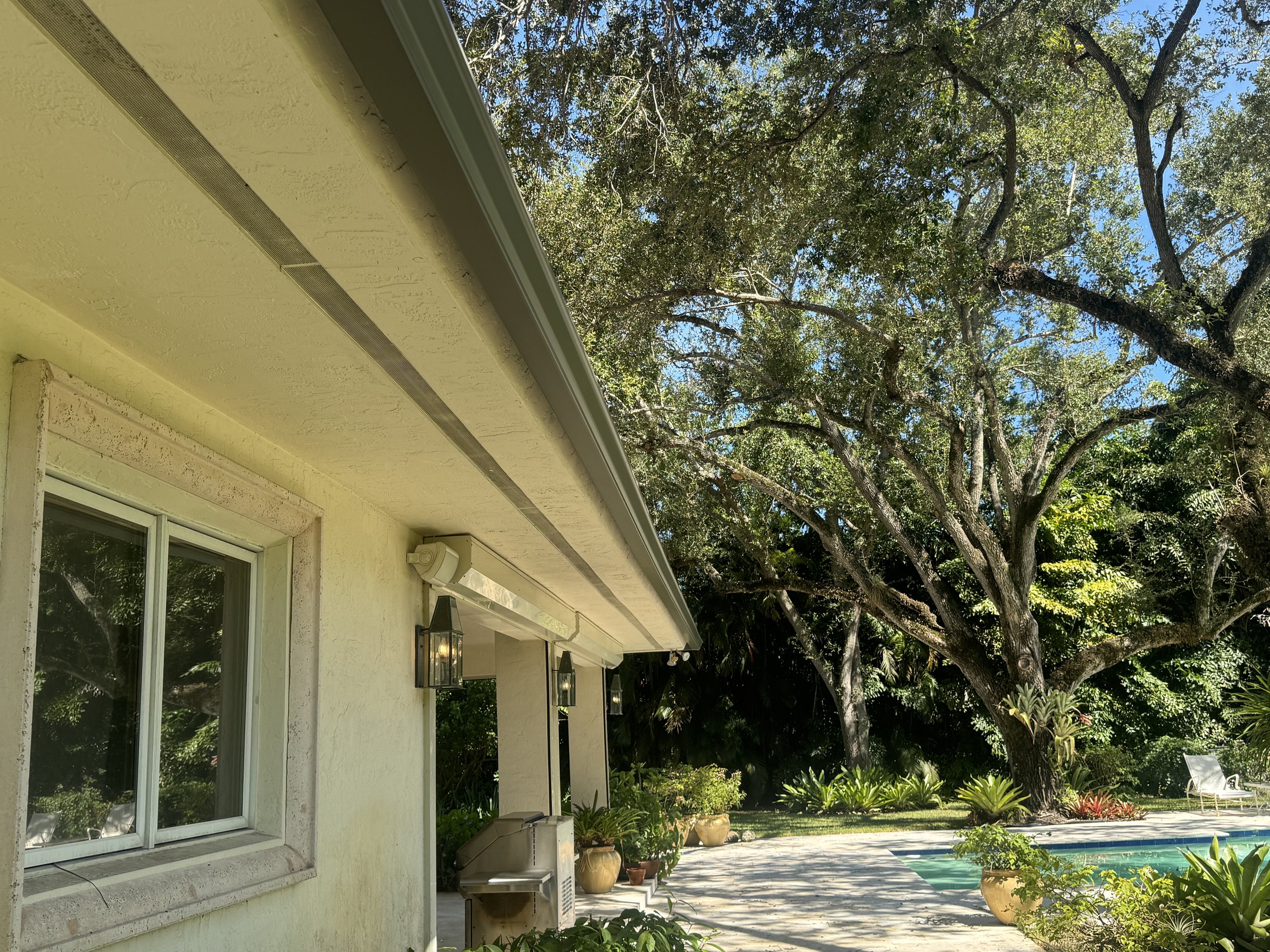 The exterior of a house rain gutters with a yellow wall, two large windows, and a porch light fixture. There is a shaded patio area with potted plants and trees, a swimming pool, and a lush garden in the background.