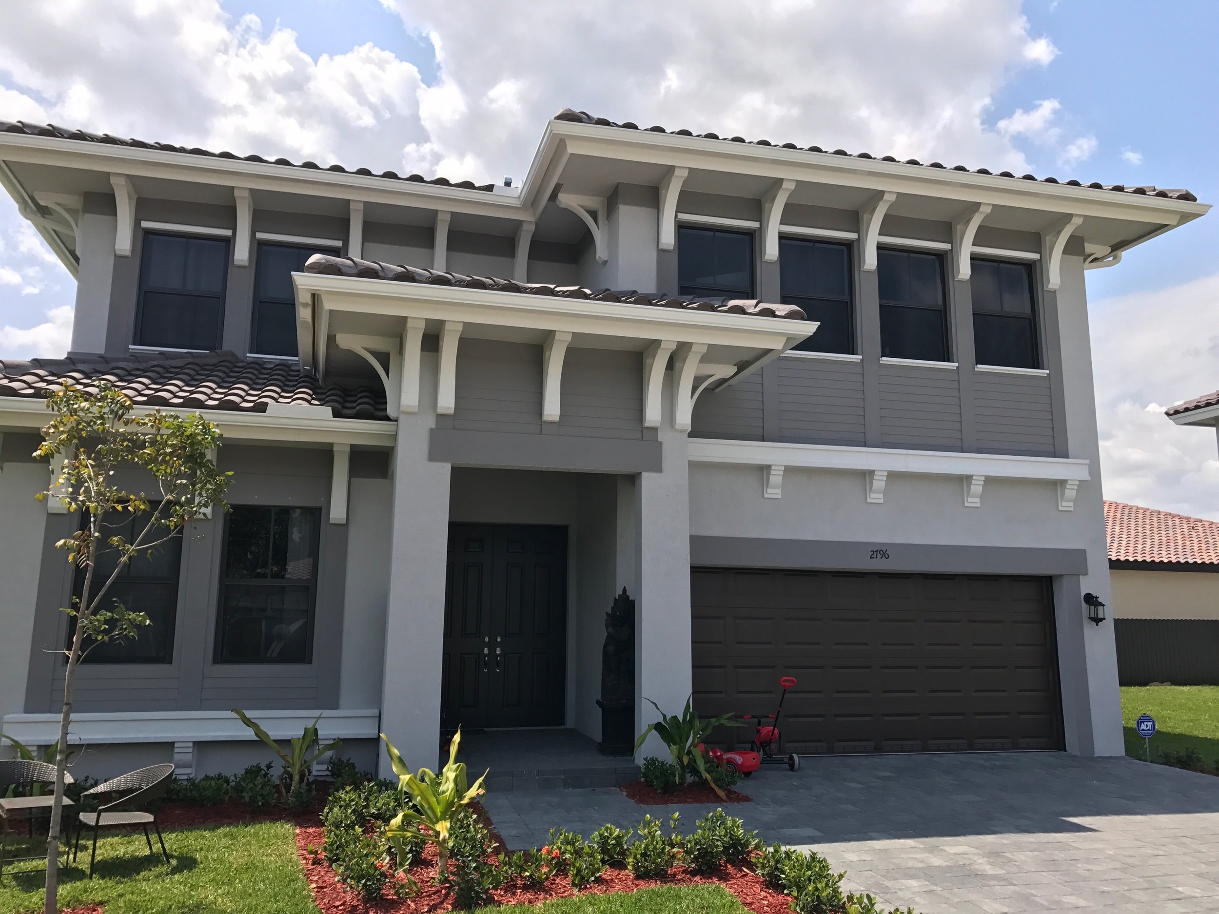 Front view of a modern, two-story house with gray exterior walls, white gutters a dark garage door, and a covered porch. The house has large windows and a tiled roof. The front yard features landscaped plants, small trees, and outdoor furniture.