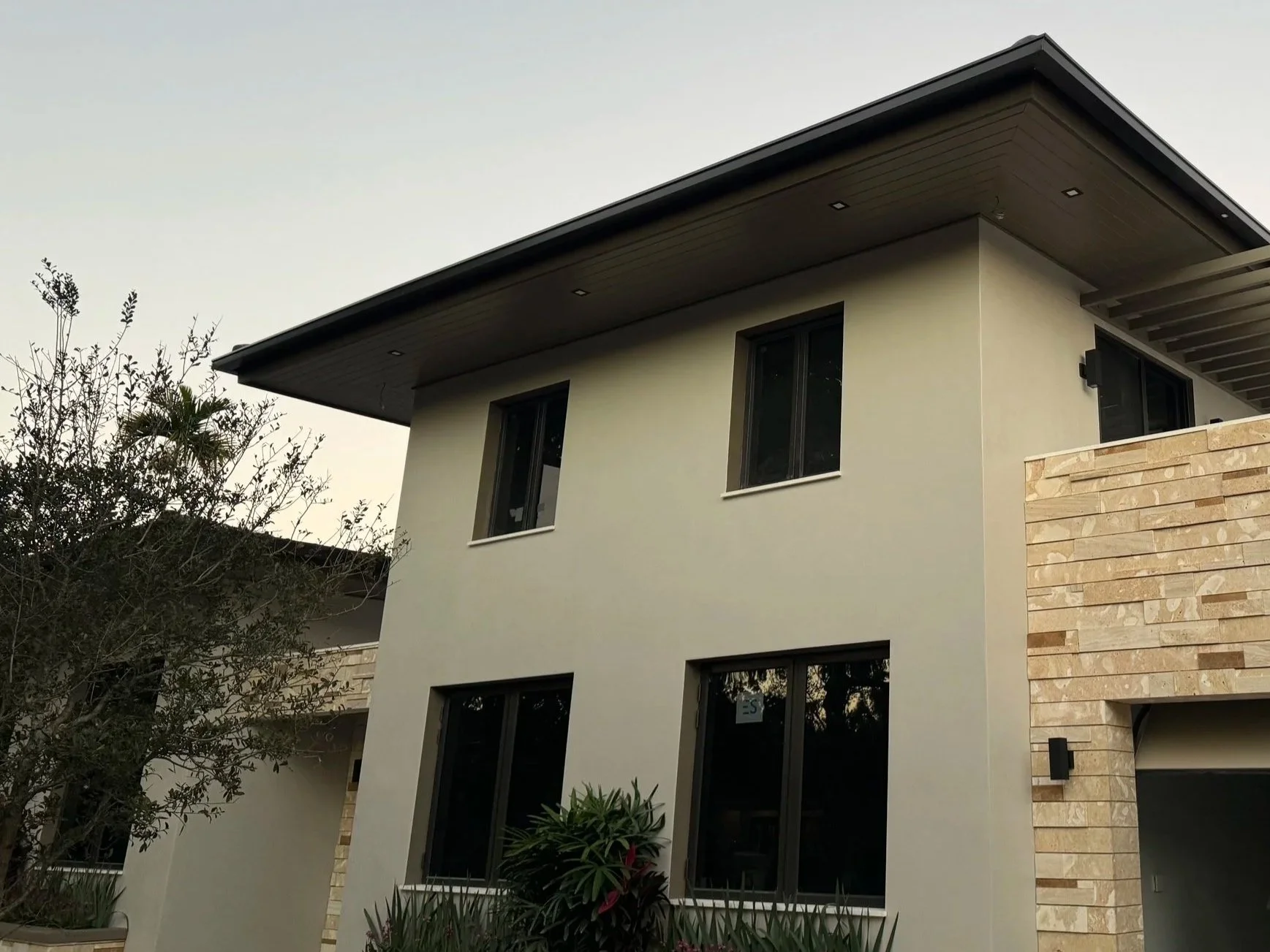 Modern two-story house with bronce gutters, beige stucco walls, large black-framed windows, and a partial stone facade, with a tree and plants in the front yard under a clear sky.