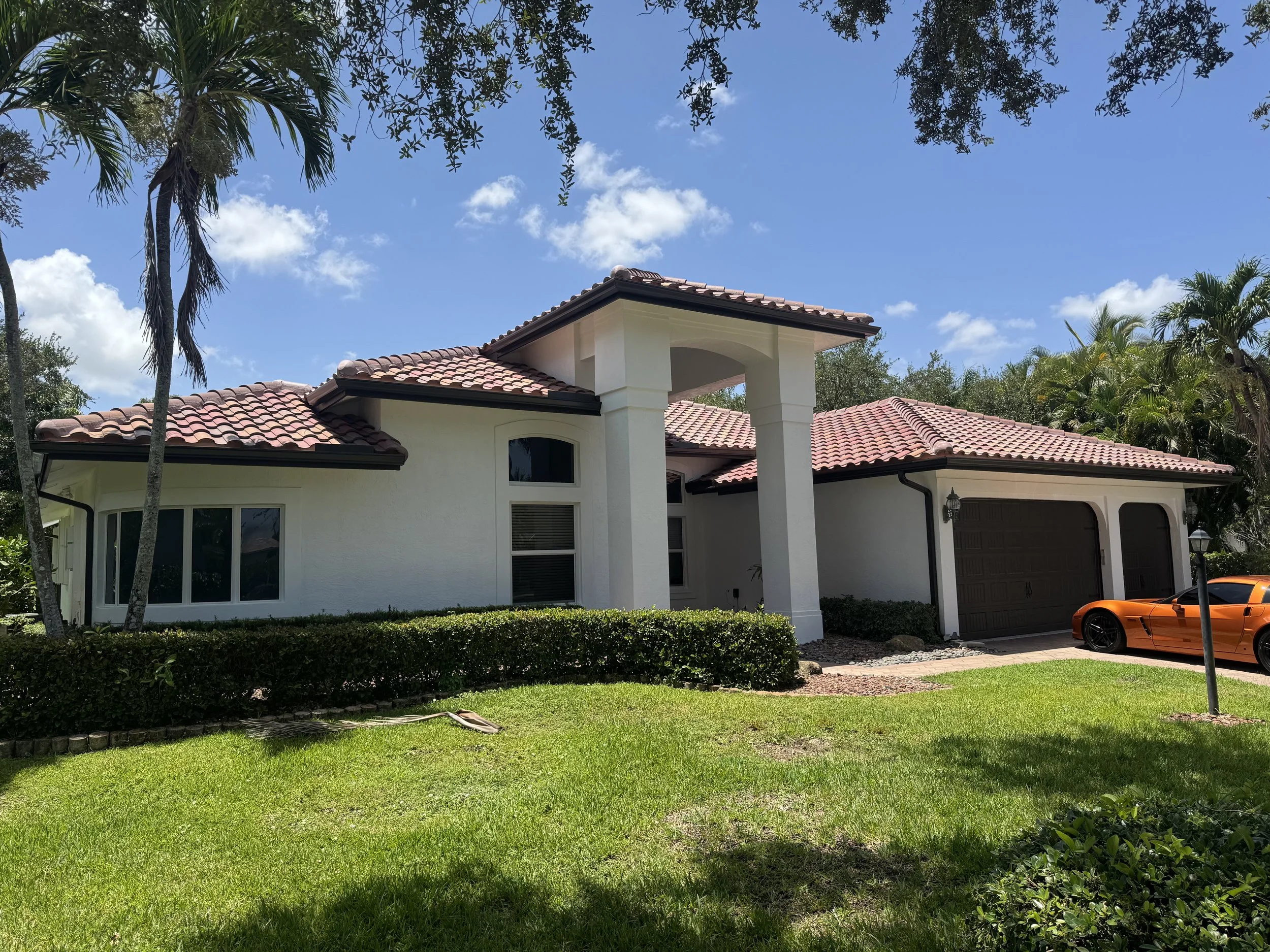 A white house with a red tiled roof and royal brown gutters, some palm trees, and a bright orange sports car parked in the driveway. The sky is blue with a few clouds.