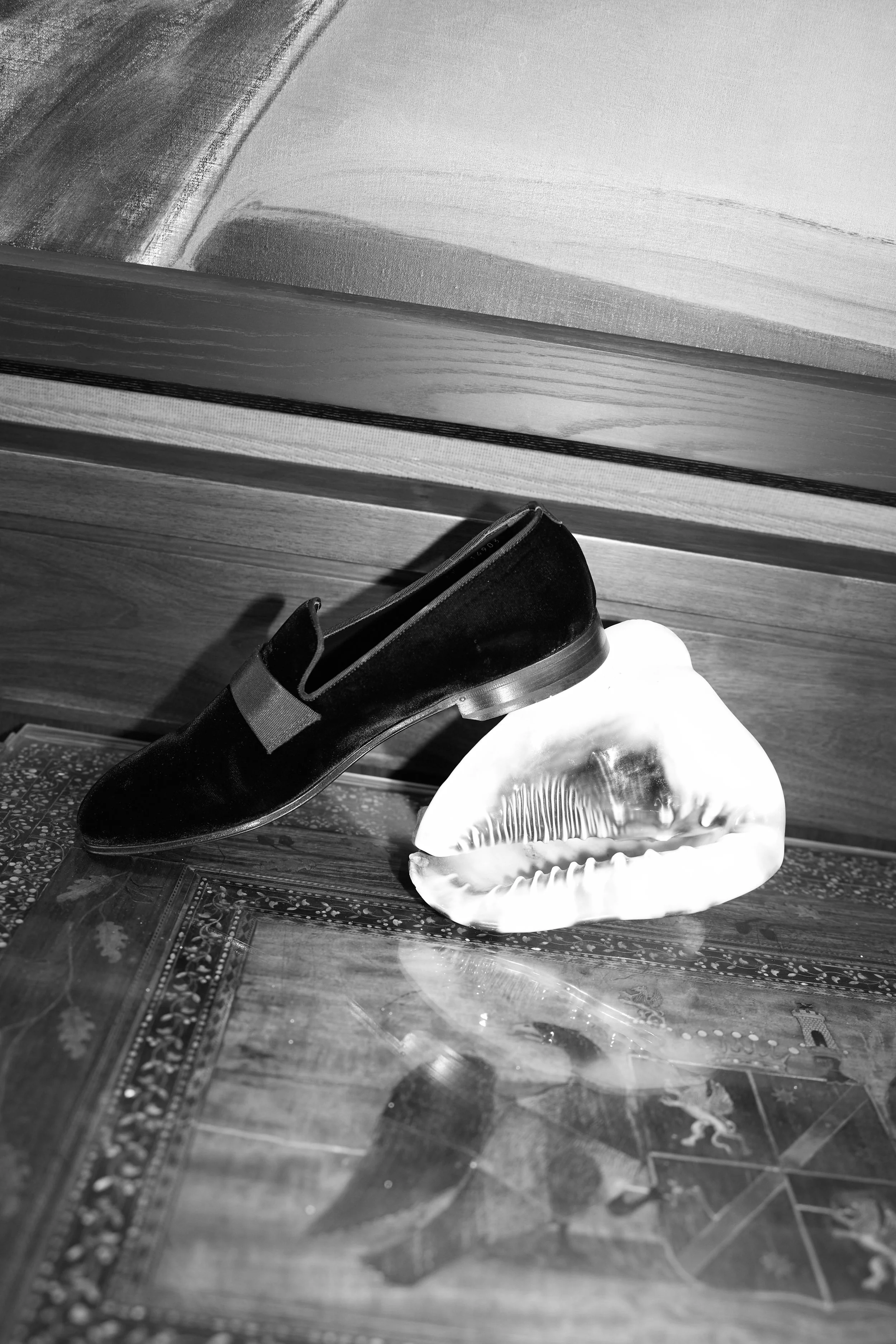 A black shoe resting on a decorative wooden surface, with a large seashell beside it. The background is wooden paneling.