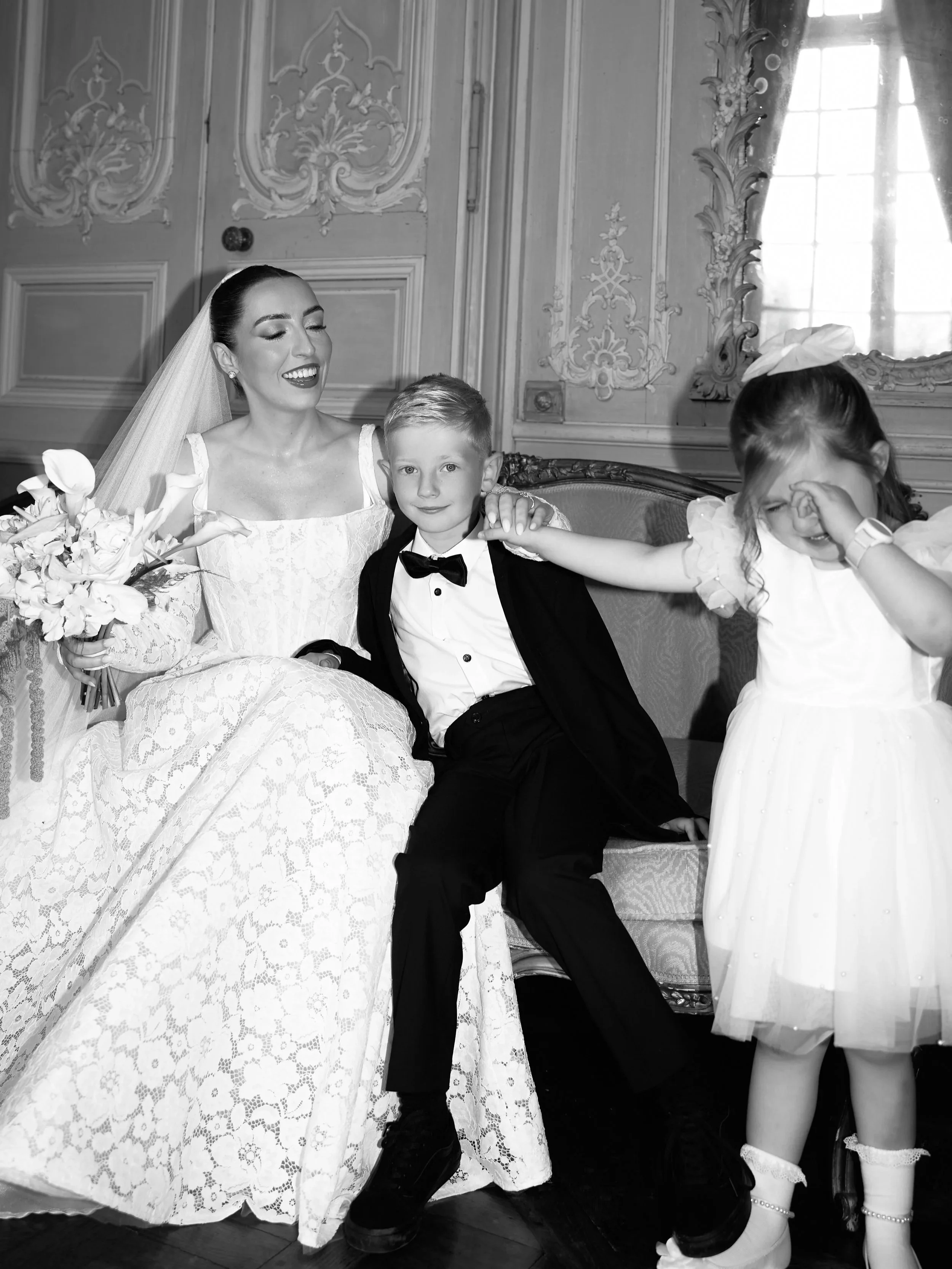 Black and white photo of a woman in a wedding dress, a young boy in a tuxedo, and a girl in a white dress indoors with ornate walls and a window with curtains. The woman is smiling, holding a bouquet. The boy is sitting between them, with her arm aro