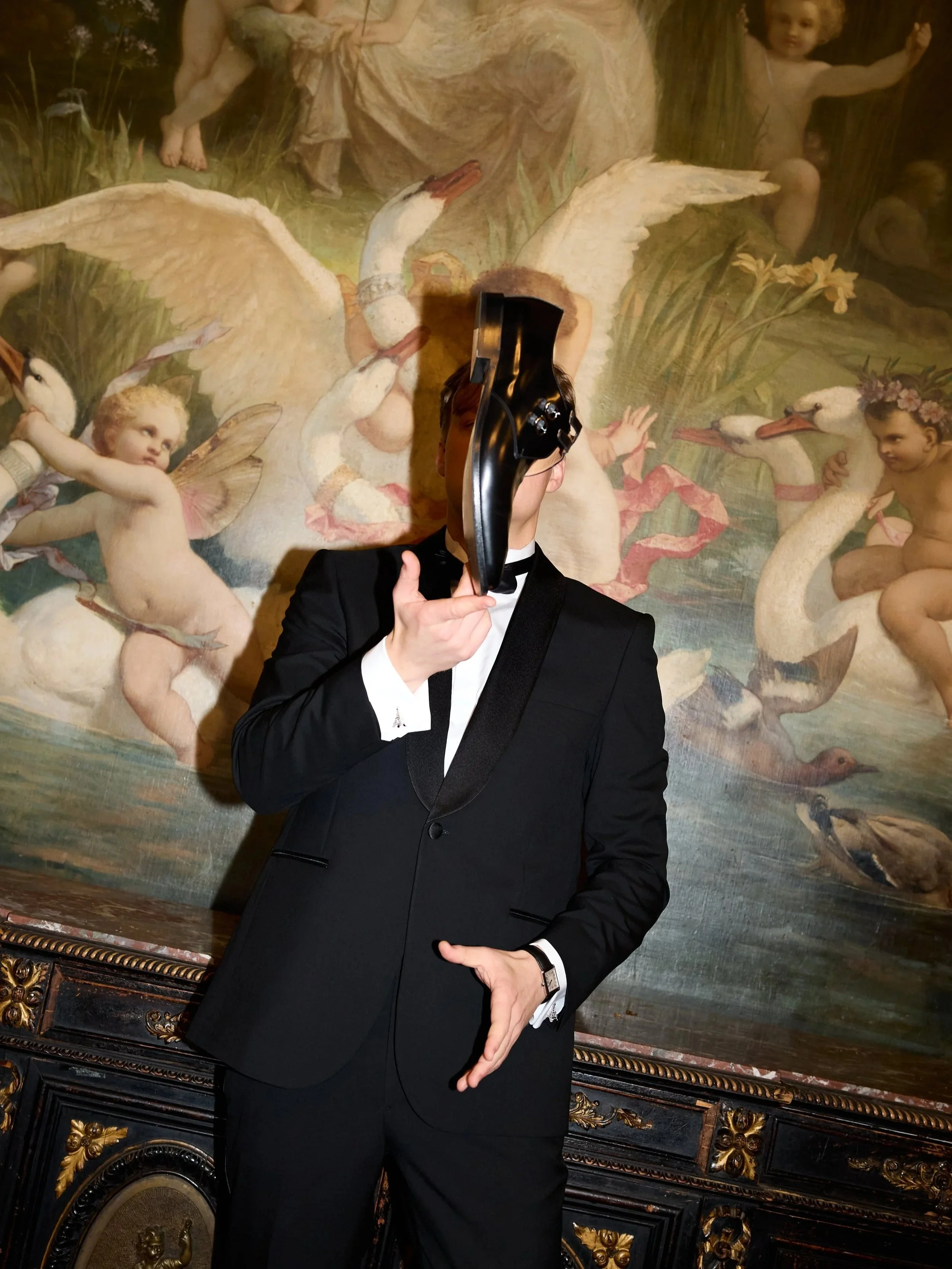 Person in a black tuxedo holding a mask in front of their face in front of a classical painting depicting mythological figures and swans.