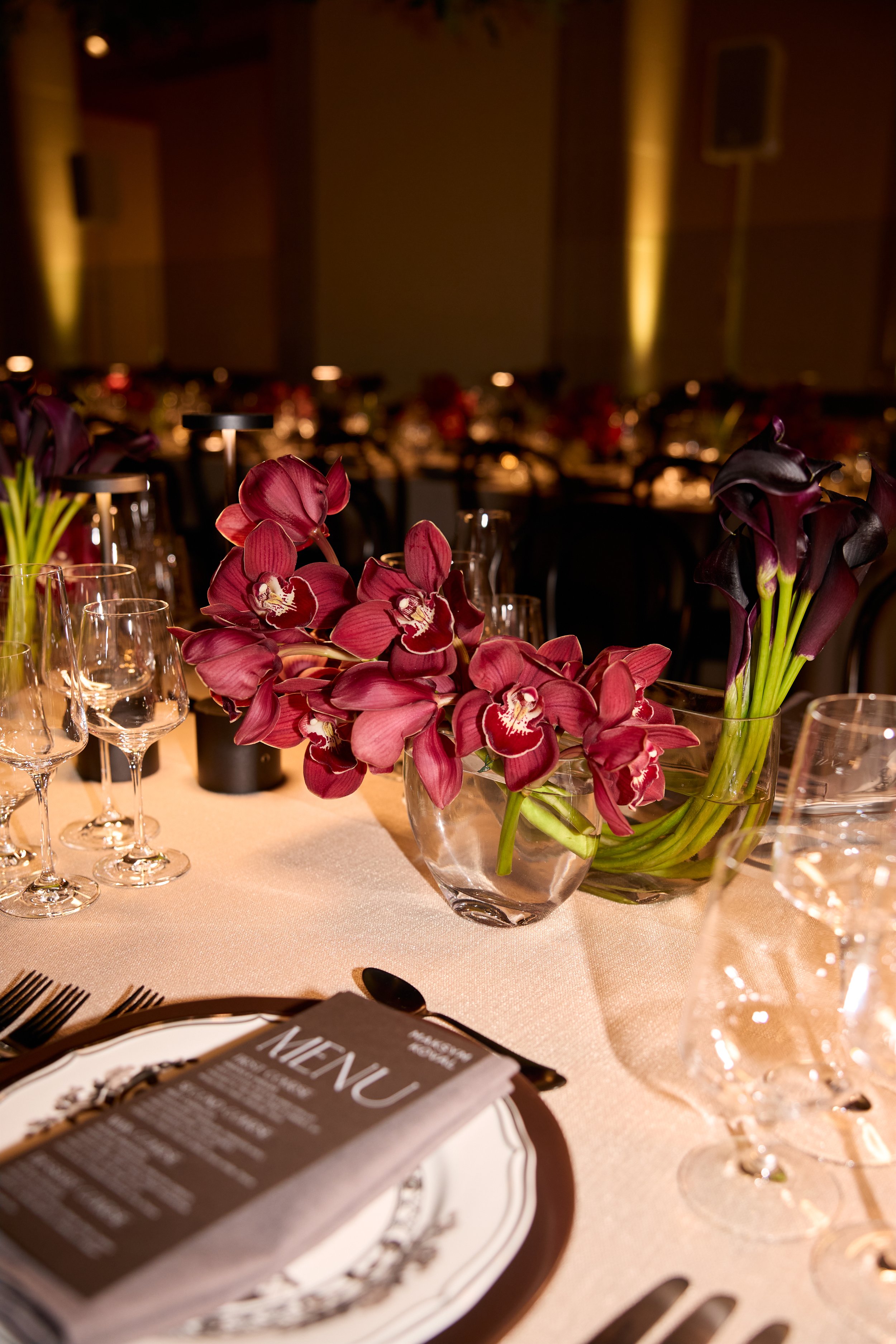 Elegant table setting with floral centerpieces featuring purple orchids and calla lilies, surrounded by empty wine glasses, a menu, and silverware.