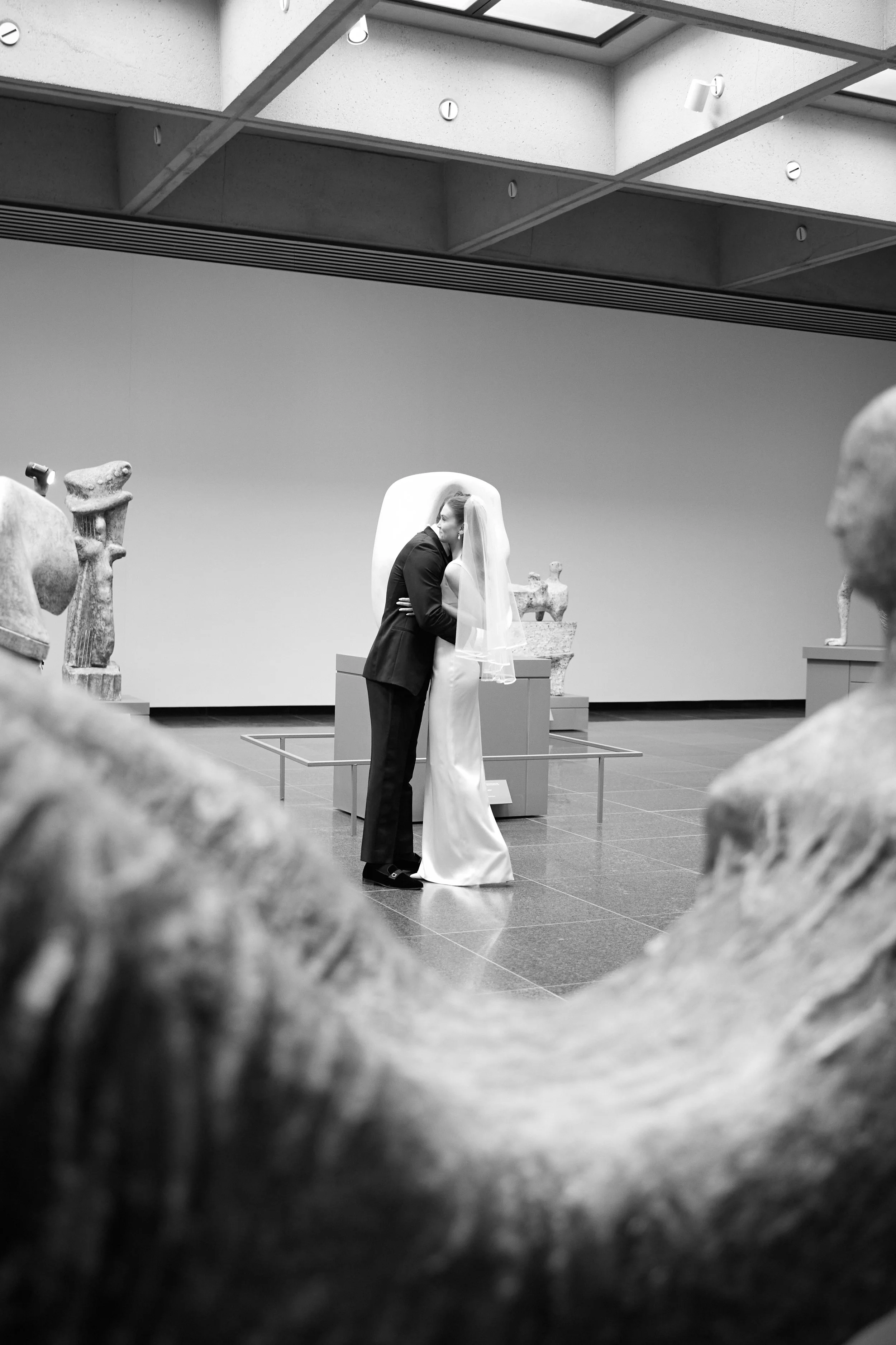 A black and white photo of a bride and groom sharing an embrace in a museum, with sculptures in the background.