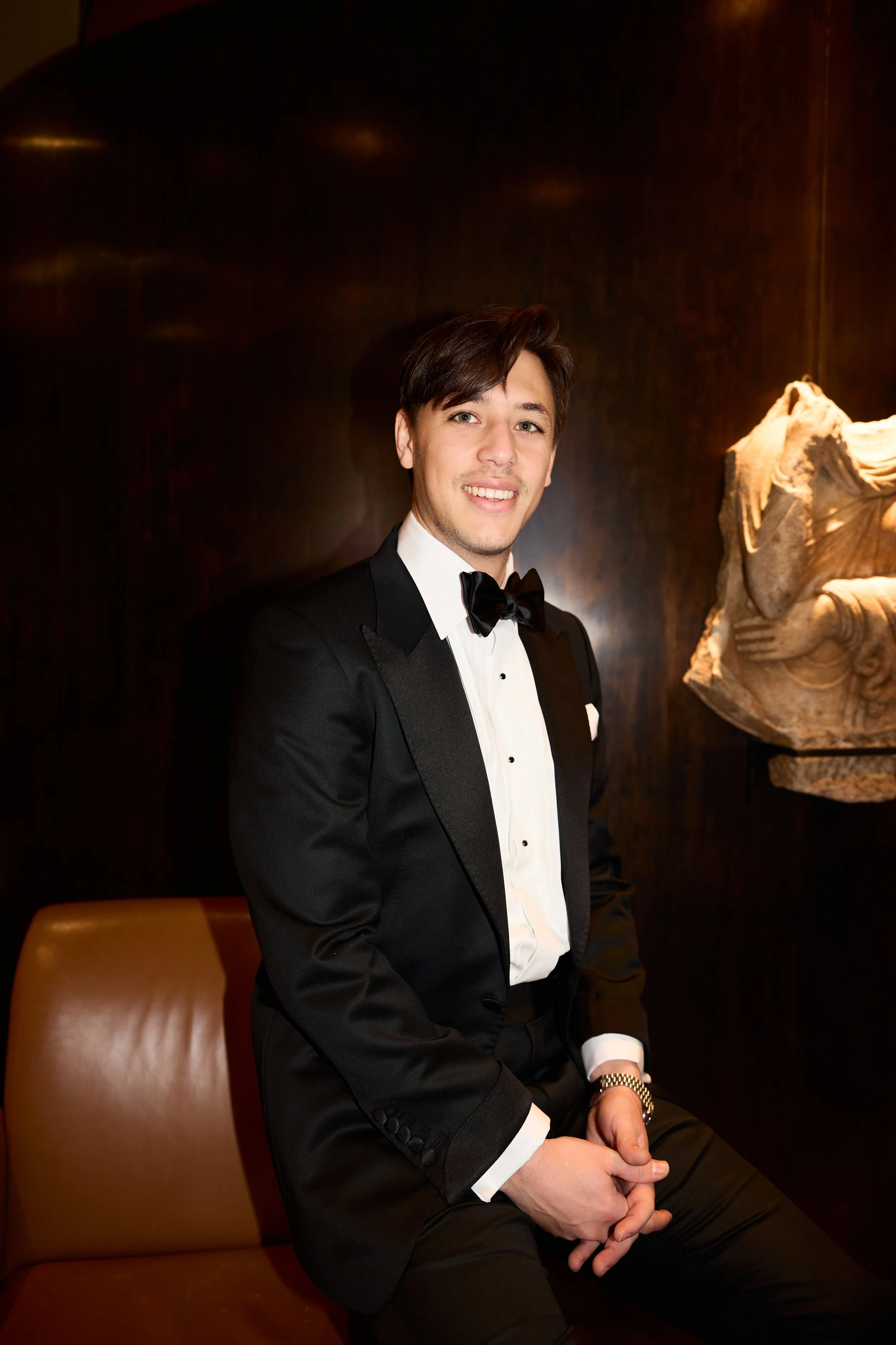 A young man in a tuxedo with a bowtie sitting on a leather chair, smiling, indoors with dark wood walls and a decorative sculpture in the background.