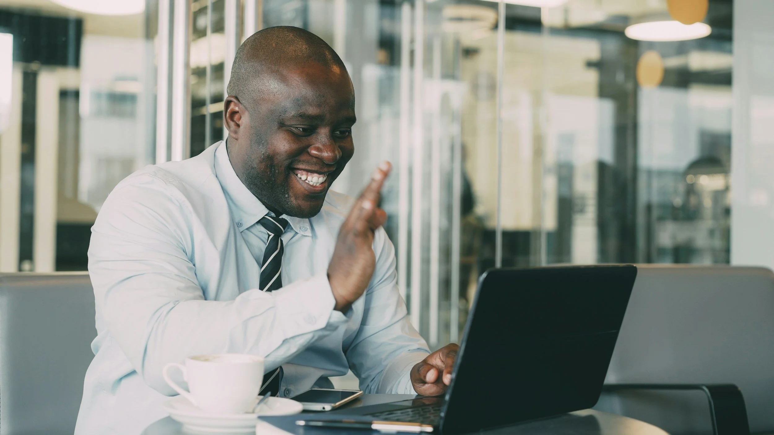 A smiling man in business attire sitting at a table with a laptop and coffee cup, appearing to be on a video call.