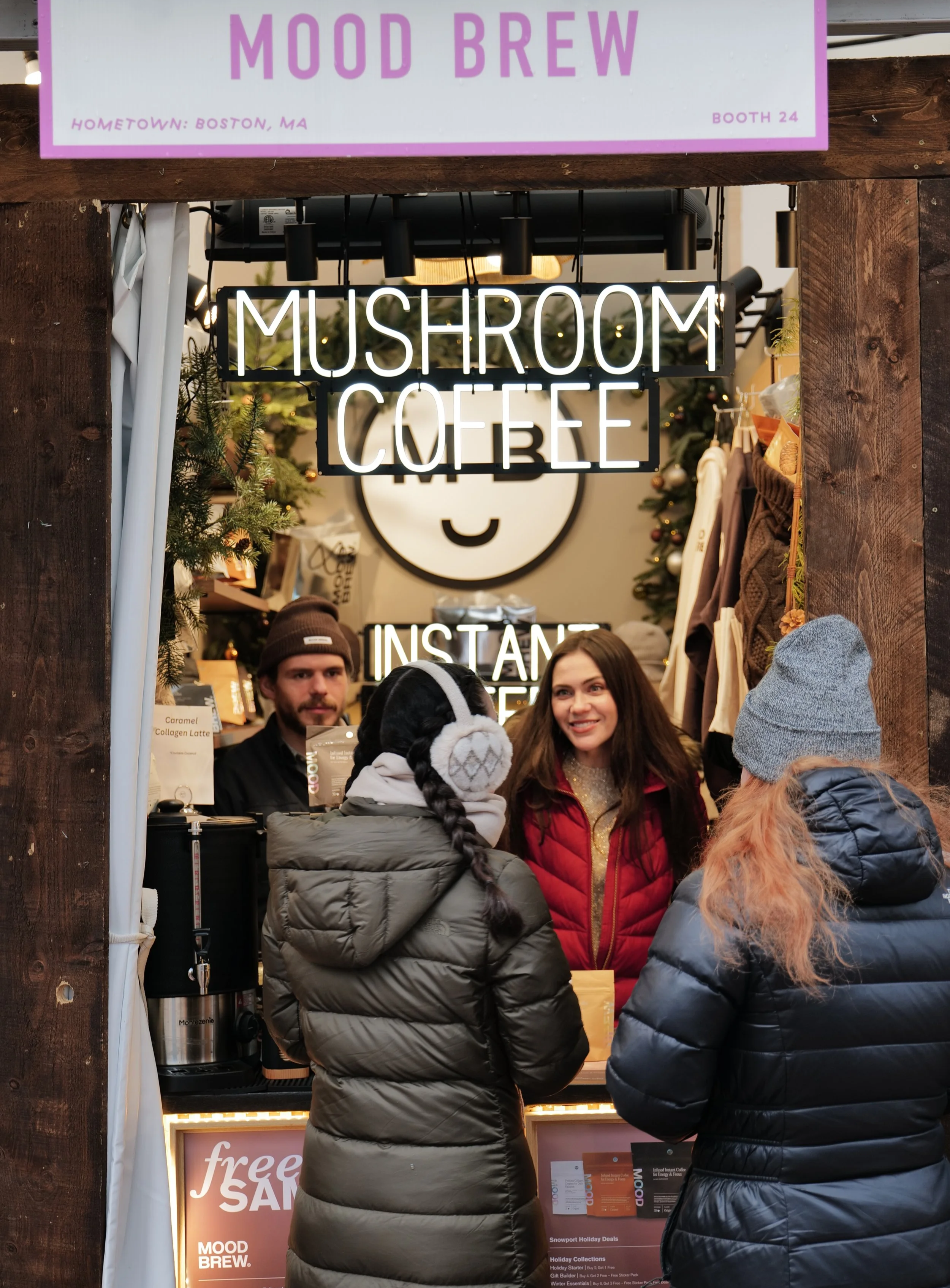 A winter scene at a holiday market stall with a neon sign reading 'Mushroom Coffee' and people ordering drinks in front. The stall has a woman with long brown hair and a man with a beard, both serving customers. The scene includes Christmas decoratio