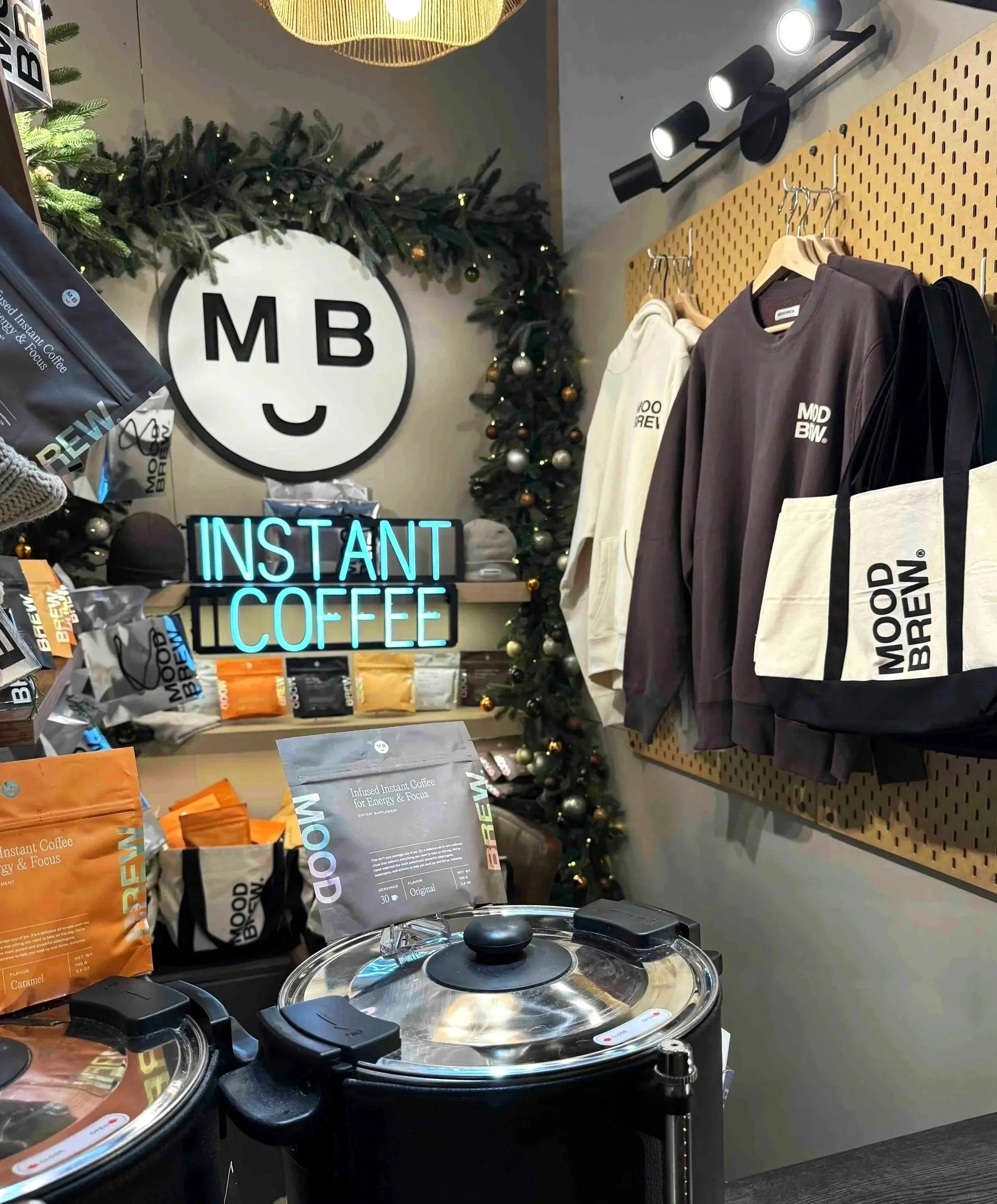 Christmas-themed display in a coffee shop with a large smiling face logo marked 'MB'. An illuminated sign reads 'INSTANT COFFEE'. Shelves feature bags of coffee and apparel with 'MOD BREW' branding. There are Christmas decorations including a wreath 