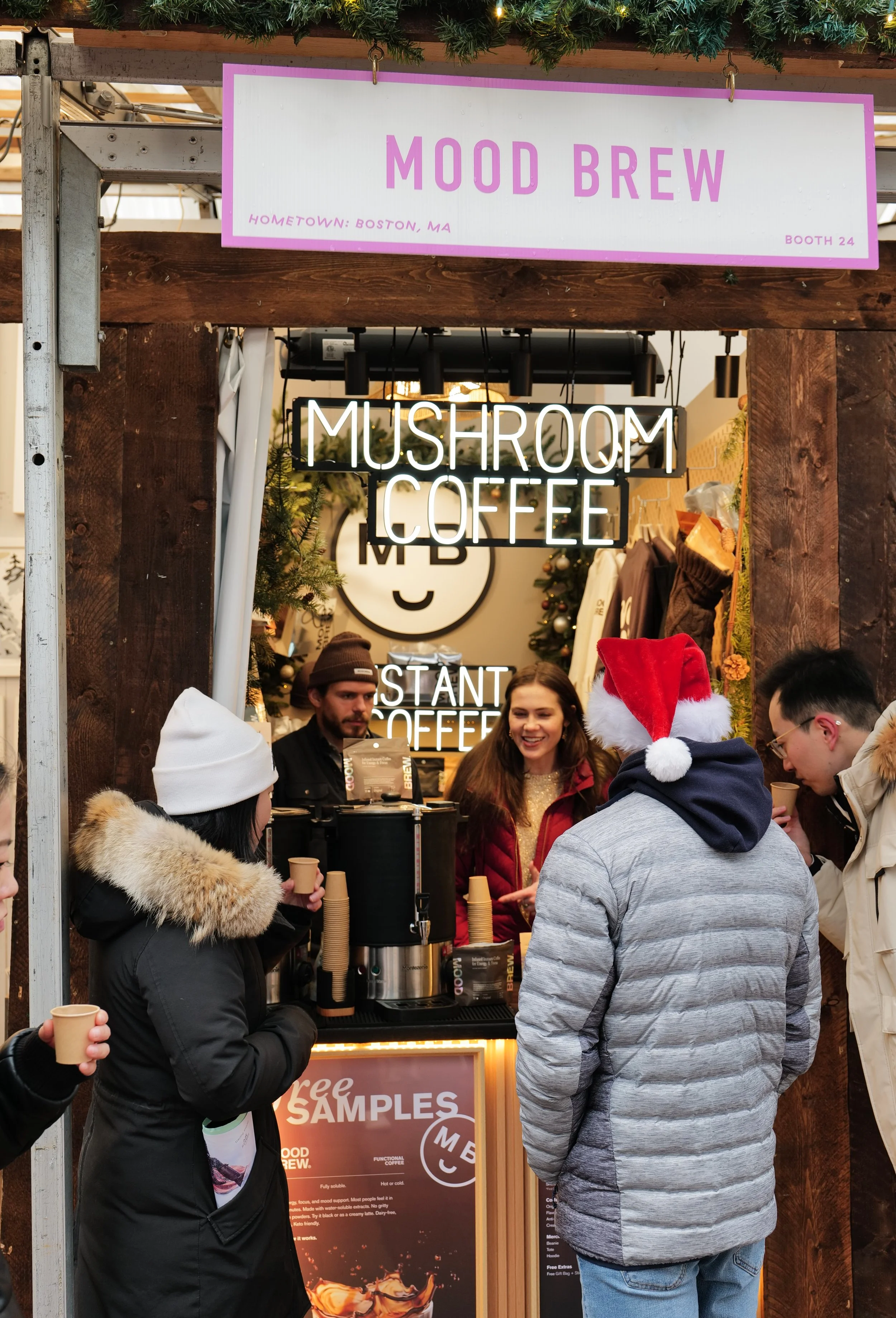 People standing at a coffee stand named Mood Brew, with signs for mushroom coffee and instant coffee. They are dressed warmly, some with holiday accessories like a Santa hat. The stand is decorated with greenery and Christmas ornaments.