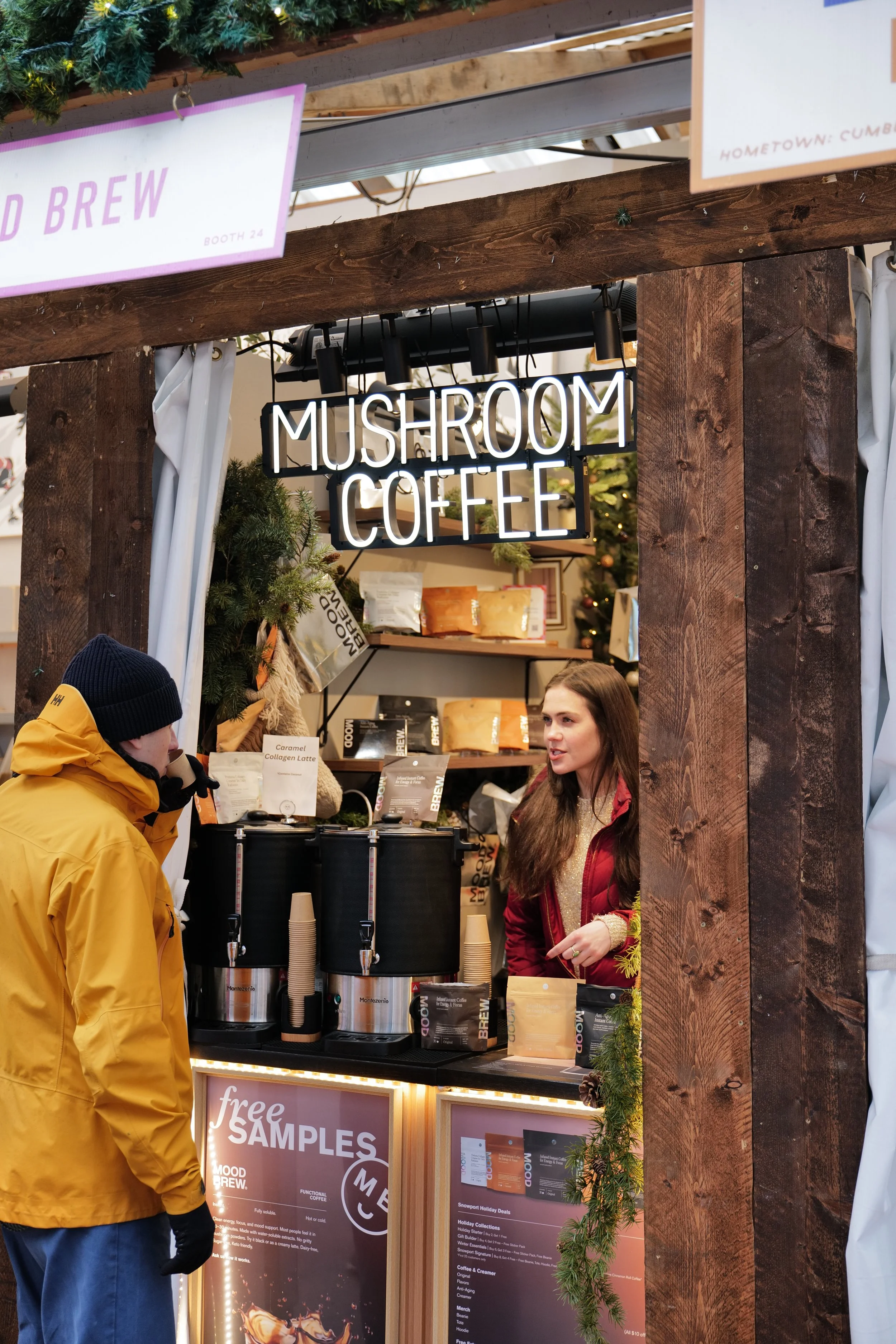 A woman serving coffee at a market stall labeled 'Mushroom Coffee.' A man in a yellow jacket and black hat stands in front of the counter. The stall decor includes pine branches, and there is a 'free samples' sign in front.