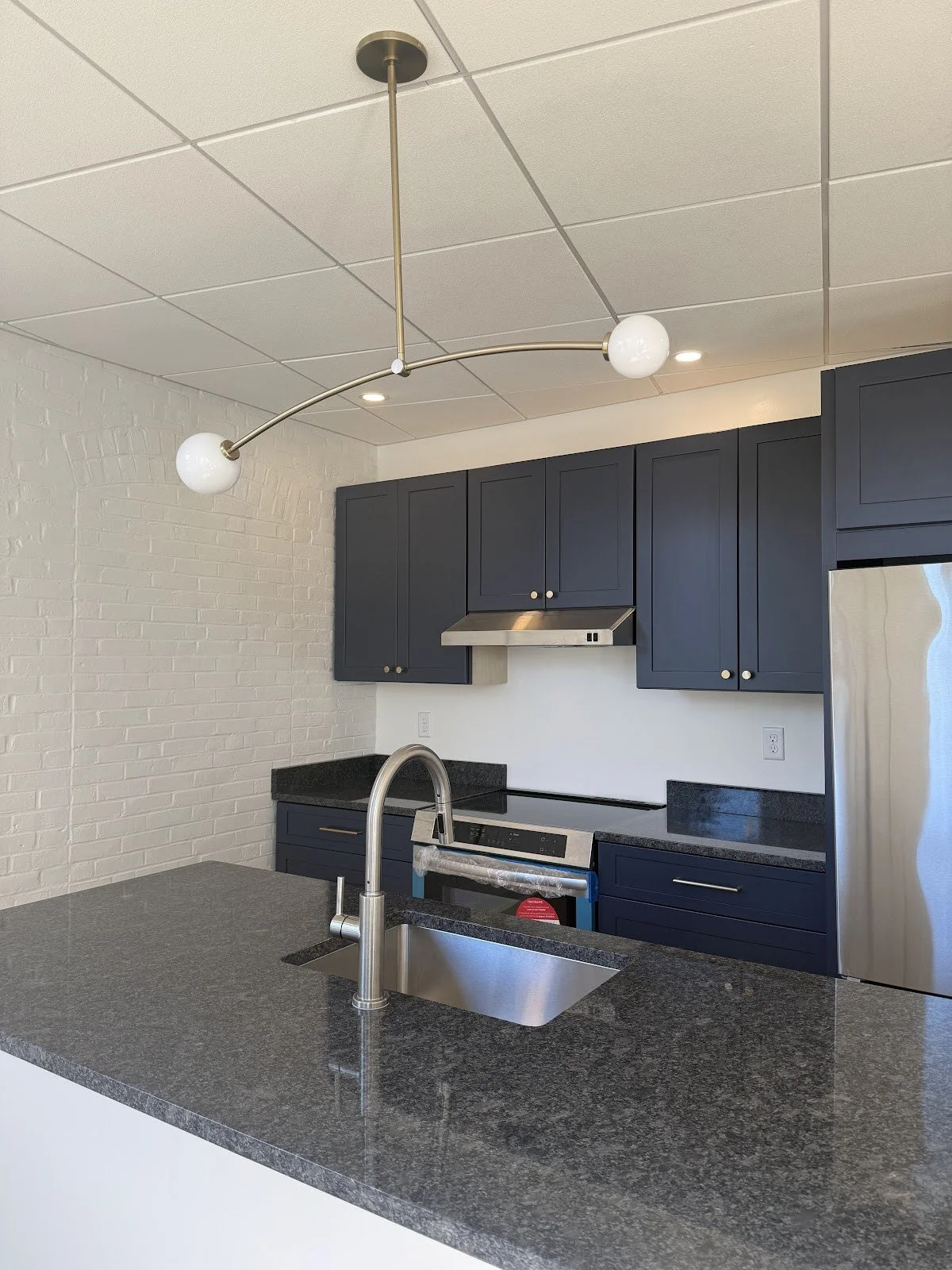 Modern kitchen with navy blue cabinets, black granite countertops, stainless steel appliances, white brick wall, and a unique ceiling light fixture with two white globes.