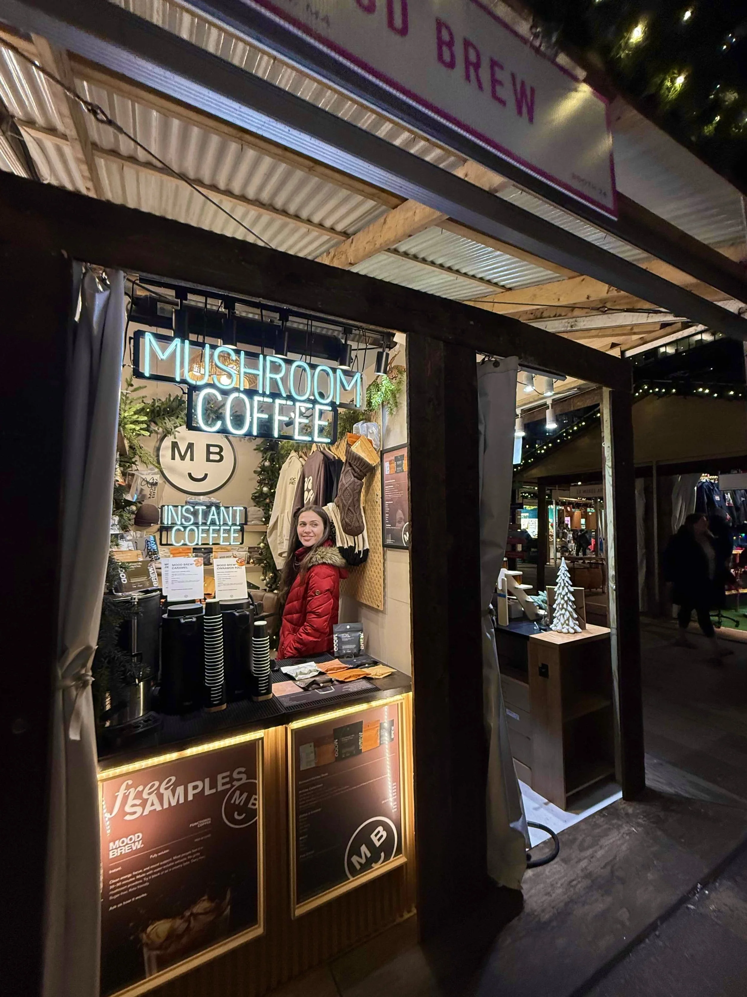 A woman in a red coat standing behind a coffee kiosk at an outdoor market during the evening. The kiosk has neon signs that say 'Mushroom Coffee' and 'Instant Coffee,' with various products and signs on the counter. The background shows other market 