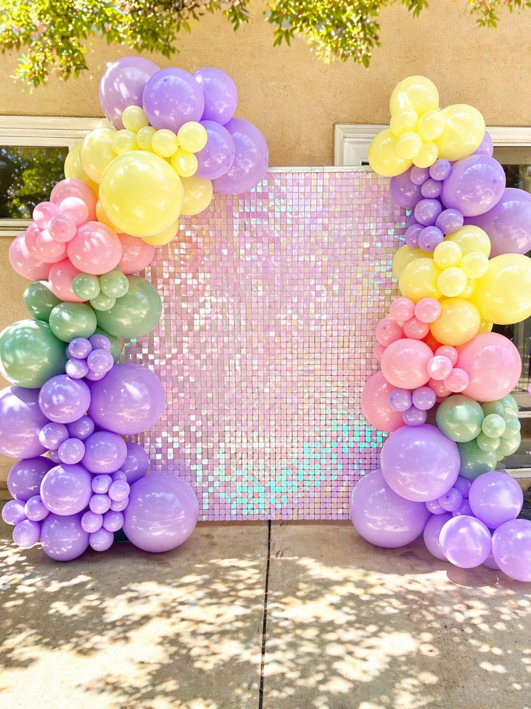 Decorative balloon arrangement with purple, pink, mint green, and white balloons in front of a pink, iridescent backdrop with the words "Autumn's Dollhouse". Cartoon character cutouts of a girl with curly hair holding a white cat and a cute mermaid-like creature with rainbow scales are also part of the display, set outdoors surrounded by trees.
