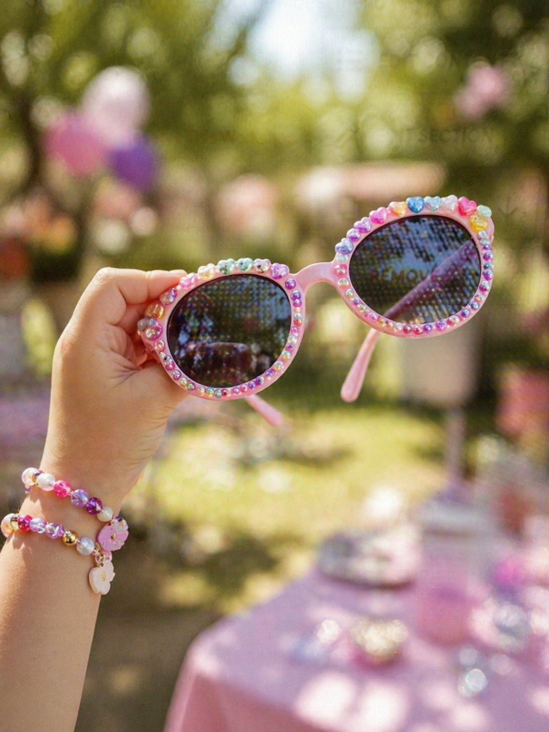 Hand holding pink sunglasses with beaded charms around the frame, with a background of other sunglasses on display. Overlay text reads: "Wearable Art Workshop: Style and decorate sunglasses and make BFF jewelry with an array of beads and charms."