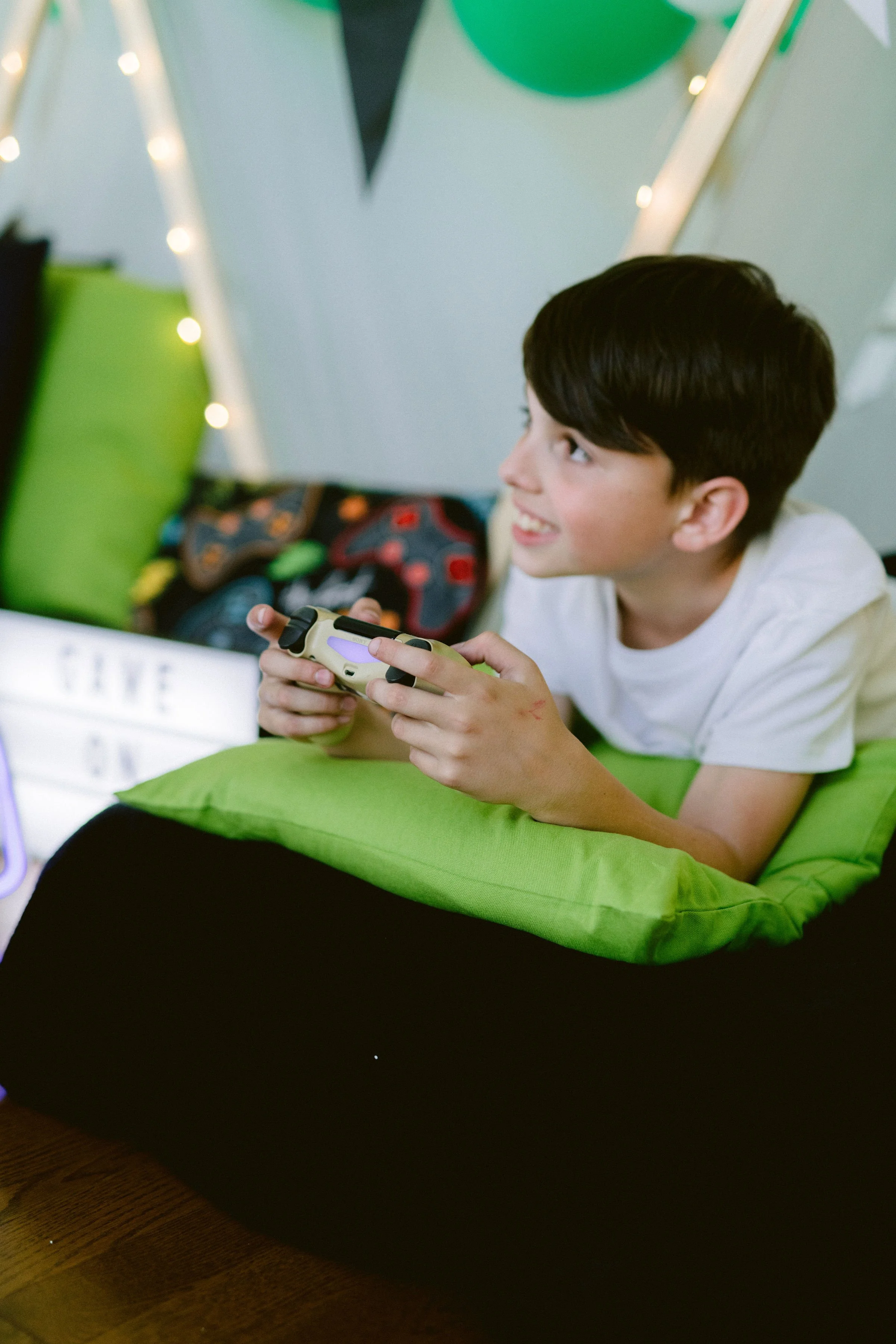A boy lying on a green pillow in bed, playing a video game with a controller. The background has a lightbox with the words "LOVE" and some decorated pillows.