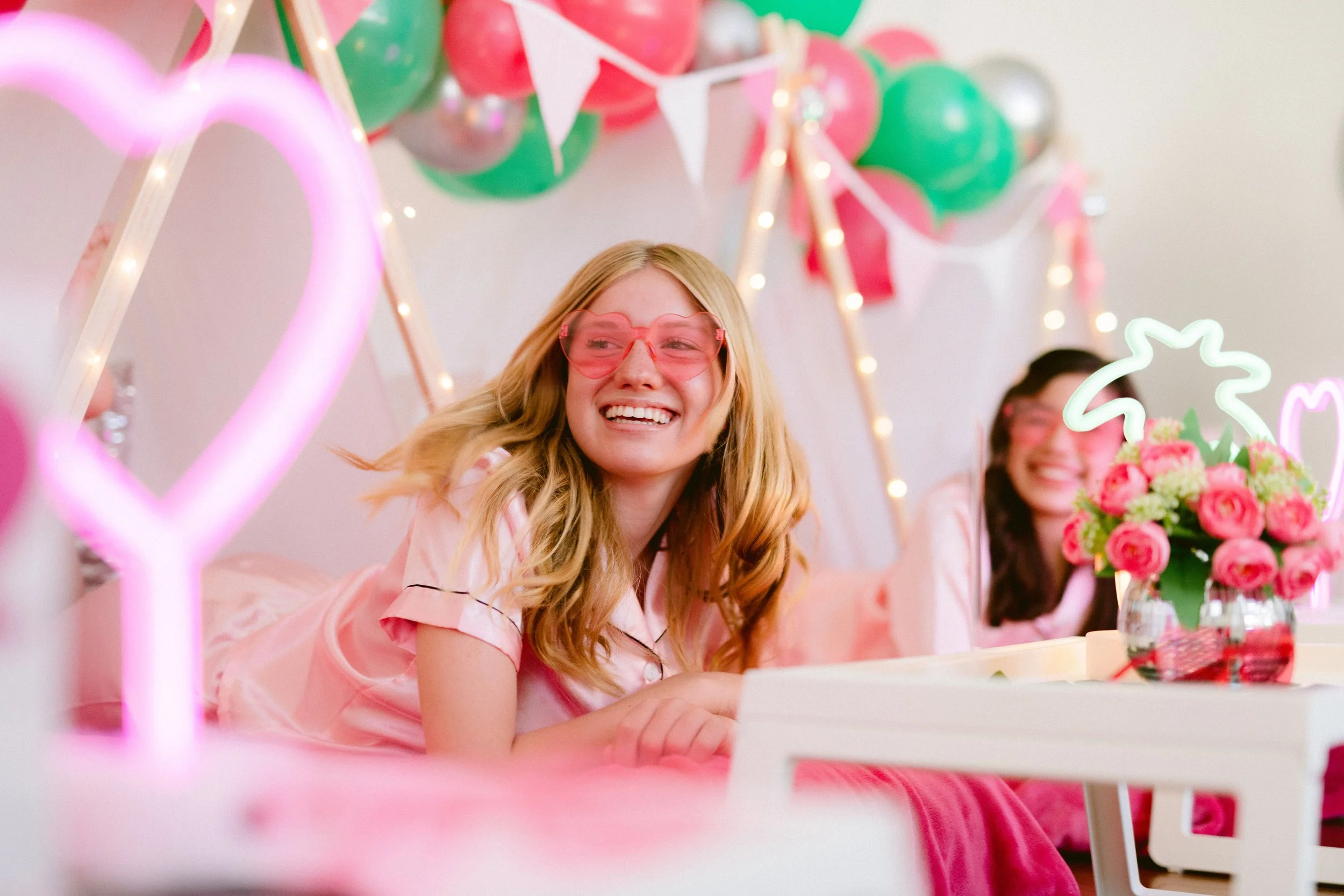 Two women in pink pajamas celebrating at a birthday party decorated with pink and green balloons, neon lights, and flowers.