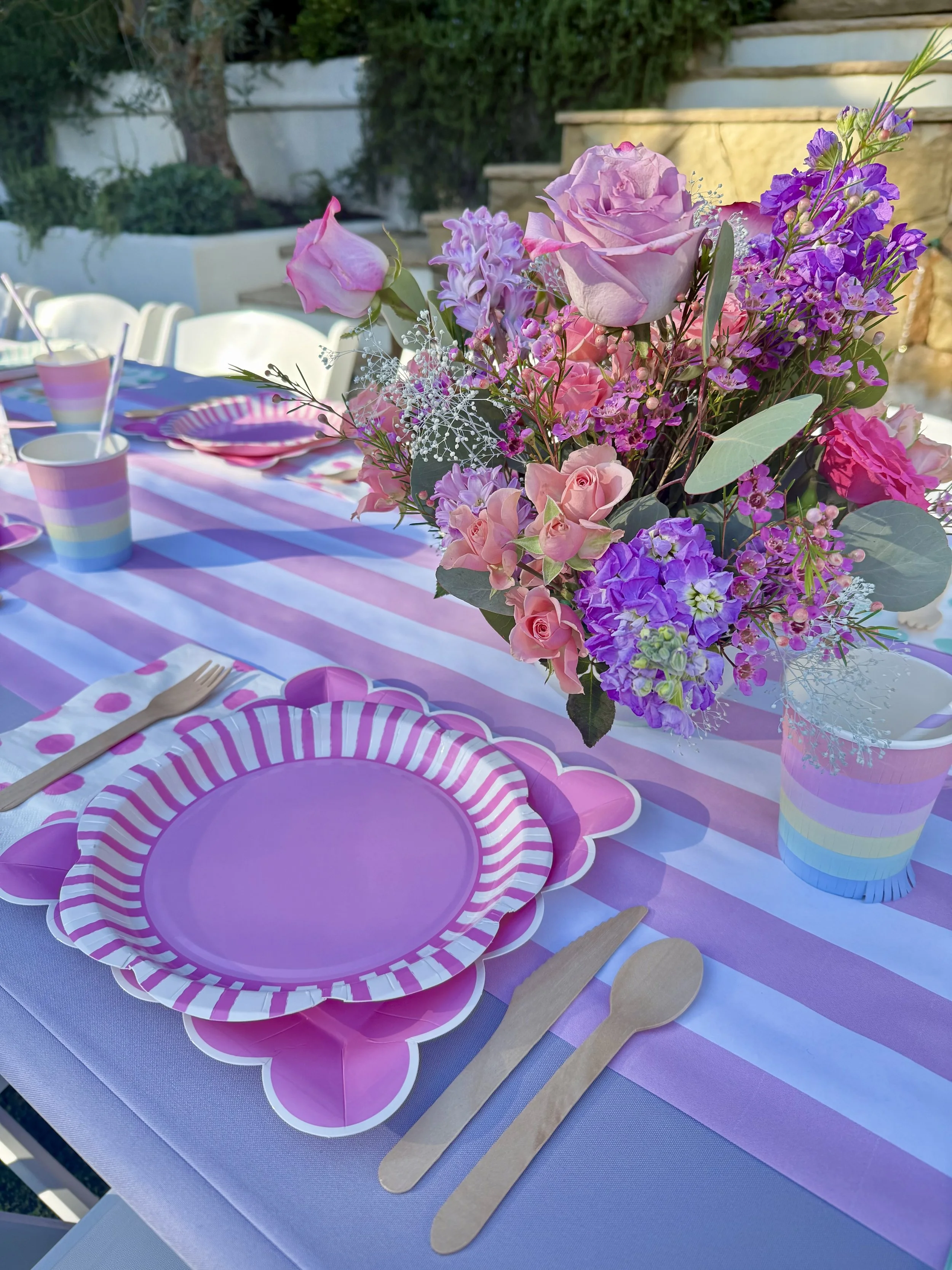 Outdoor party setup featuring a long table with pink tablecloths, decorated with strawberry-themed plates, cups, and napkins, surrounded by white chairs. At the end of the table, there is a backdrop made of pink and red balloons shaped like strawberries, situated among green trees.