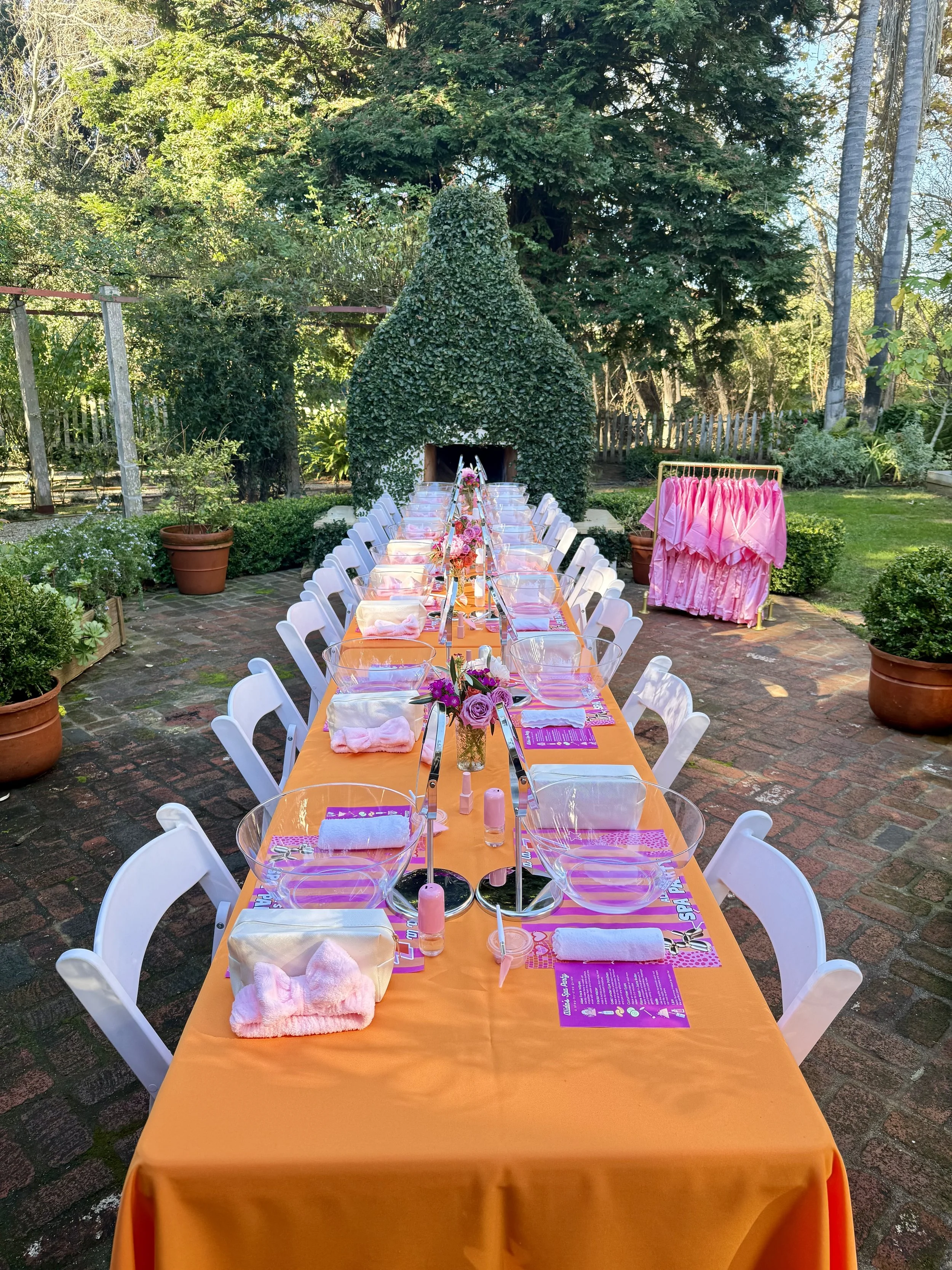 Outdoor dining table decorated with an orange tablecloth, set with clear bowls, pink napkins, and floral centerpieces, arranged for a party or event in a garden with trees and potted plants in the background.