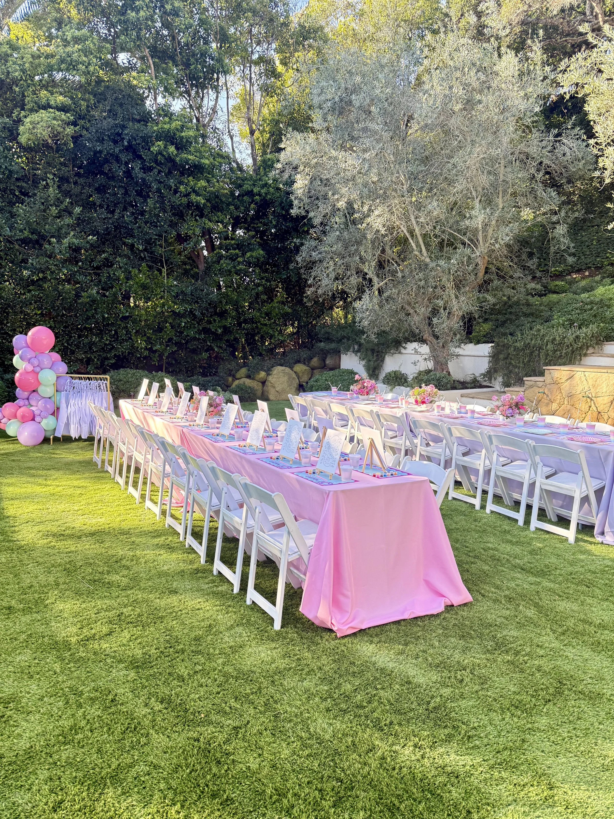 Outdoor party setup with long pink tablecloth-covered tables, white chairs, pink and purple balloons, floral centerpieces, and place settings, set in a garden with green grass and trees.