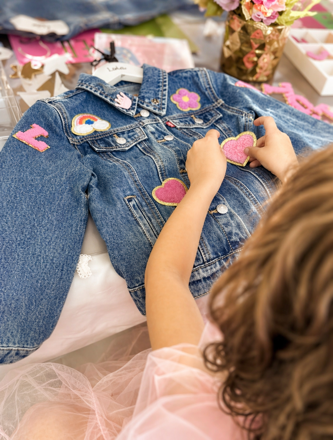 Child placing pink and yellow heart patches on a denim jacket decorated with other patches like rainbow and flower.