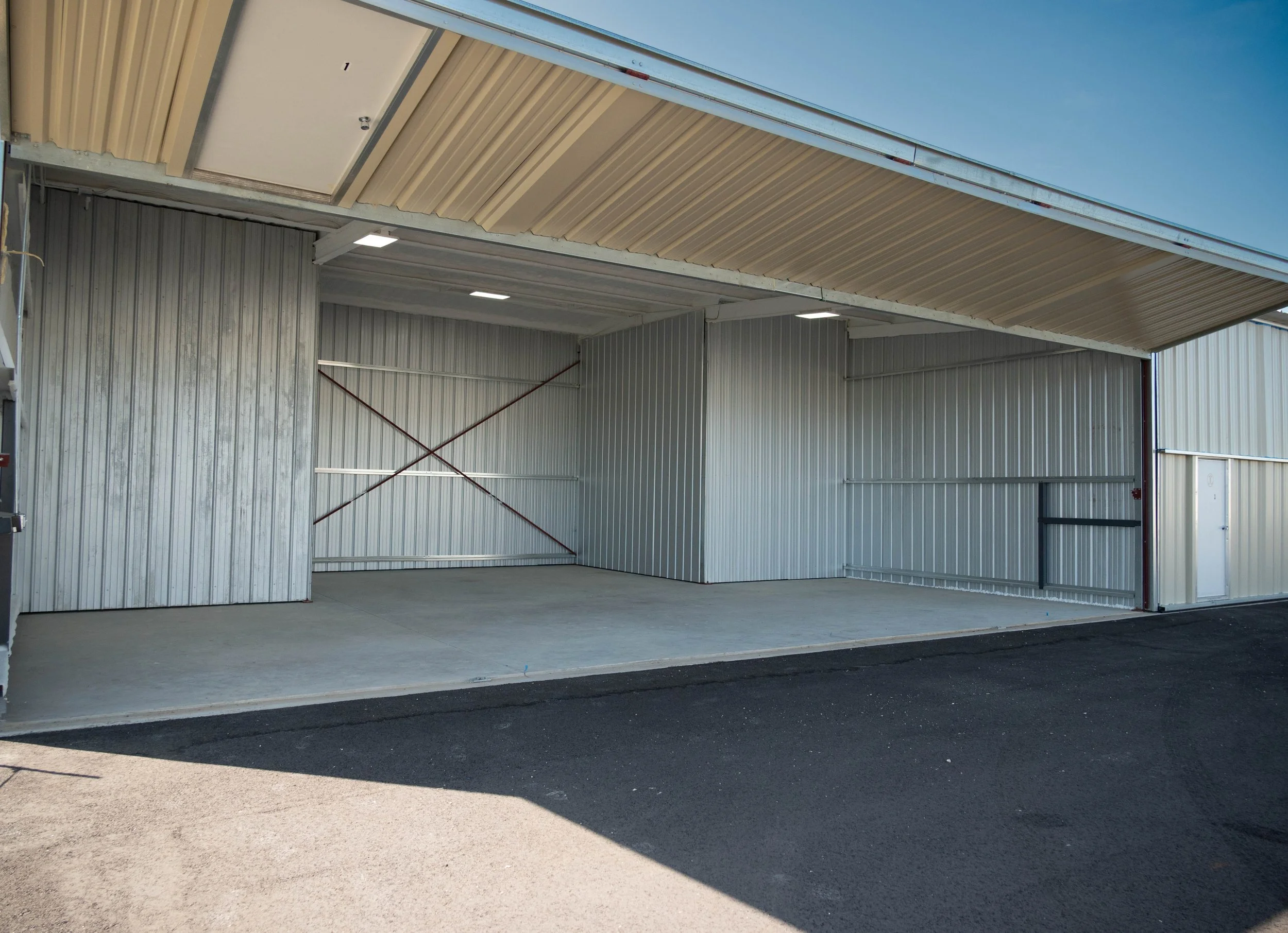 Open metal storage unit with concrete floor, metal walls, and partially open sliding door, with a black paved surface outside.