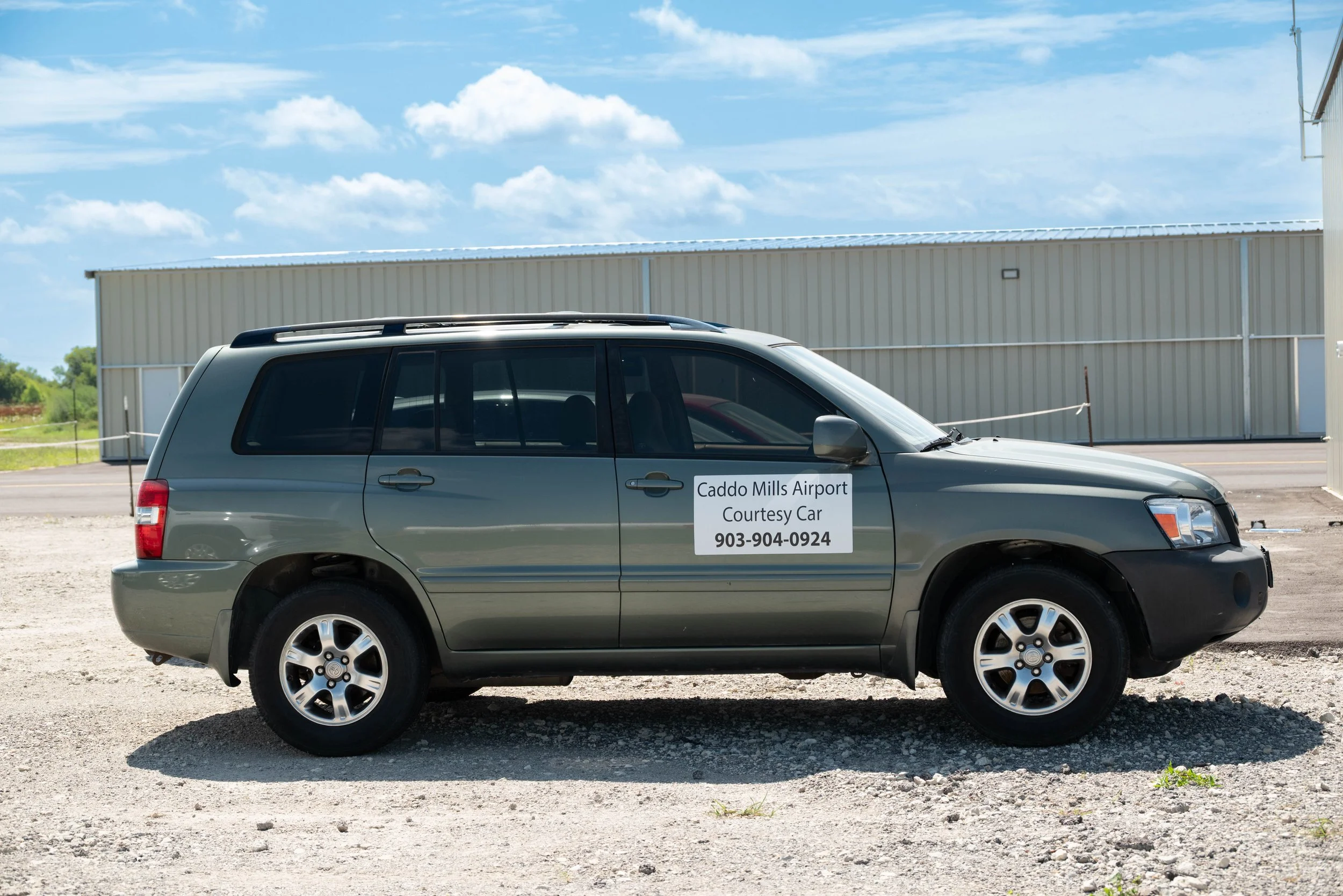 Gray SUV parked on a gravel lot under a blue sky with clouds, with a sign on the side that reads 'Caddo Mills Airport Courtesy Car 903-904-0924'.
