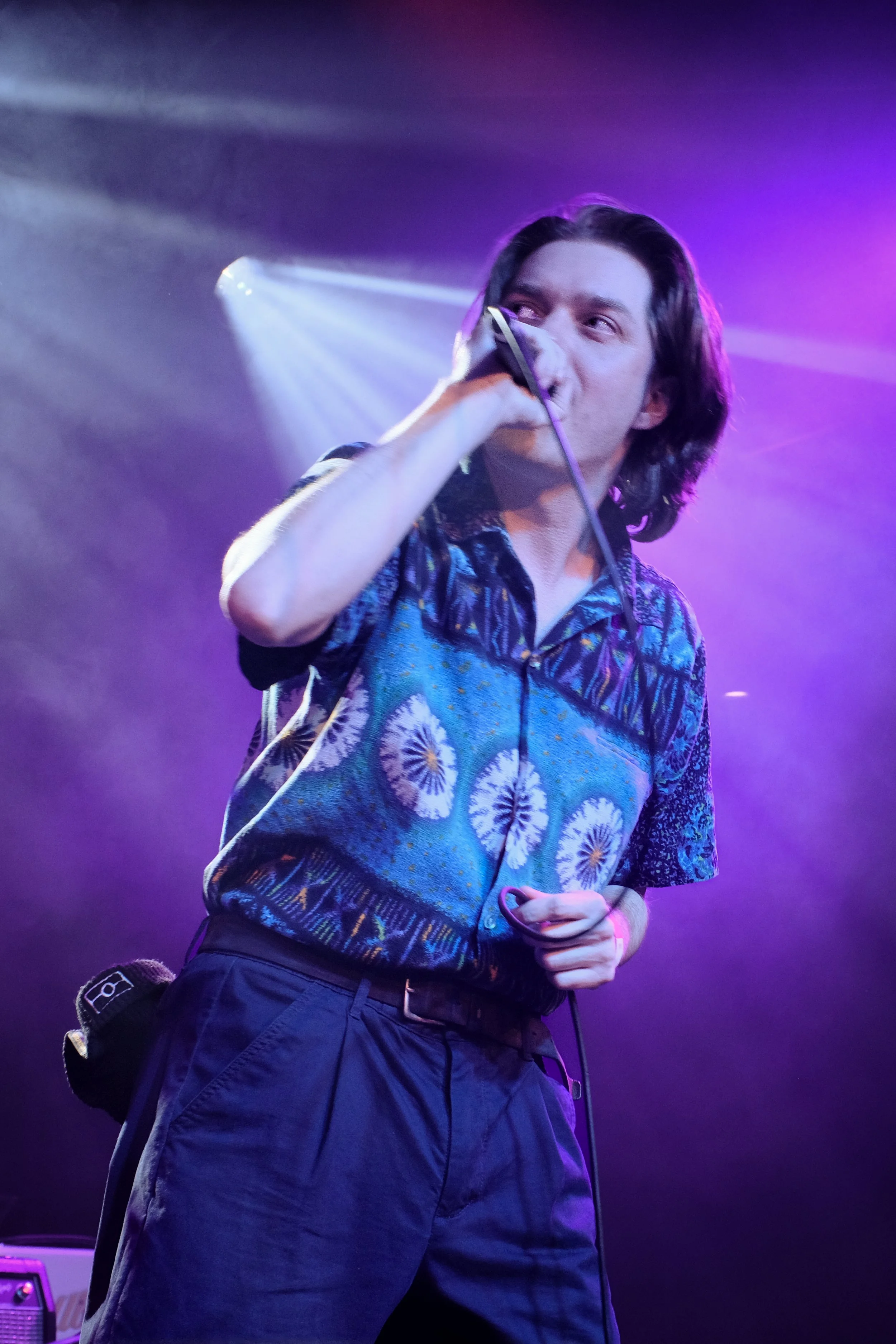 A male singer with shoulder-length dark hair sings into a microphone on stage, illuminated by purple and blue stage lights, wearing a colorful patterned shirt and dark pants.