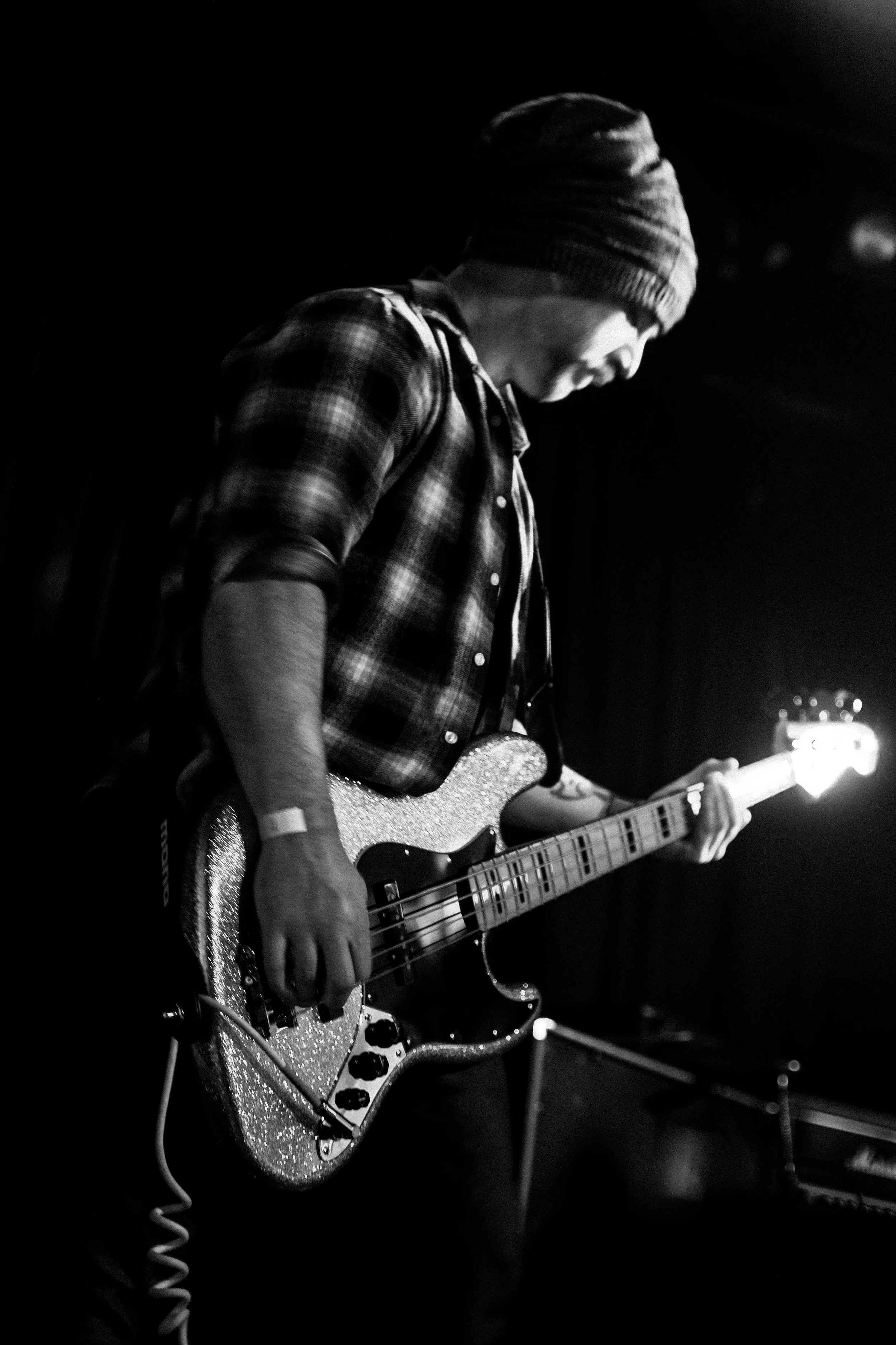 A person wearing a beanie and checkered shirt playing an electric guitar on stage in black and white.
