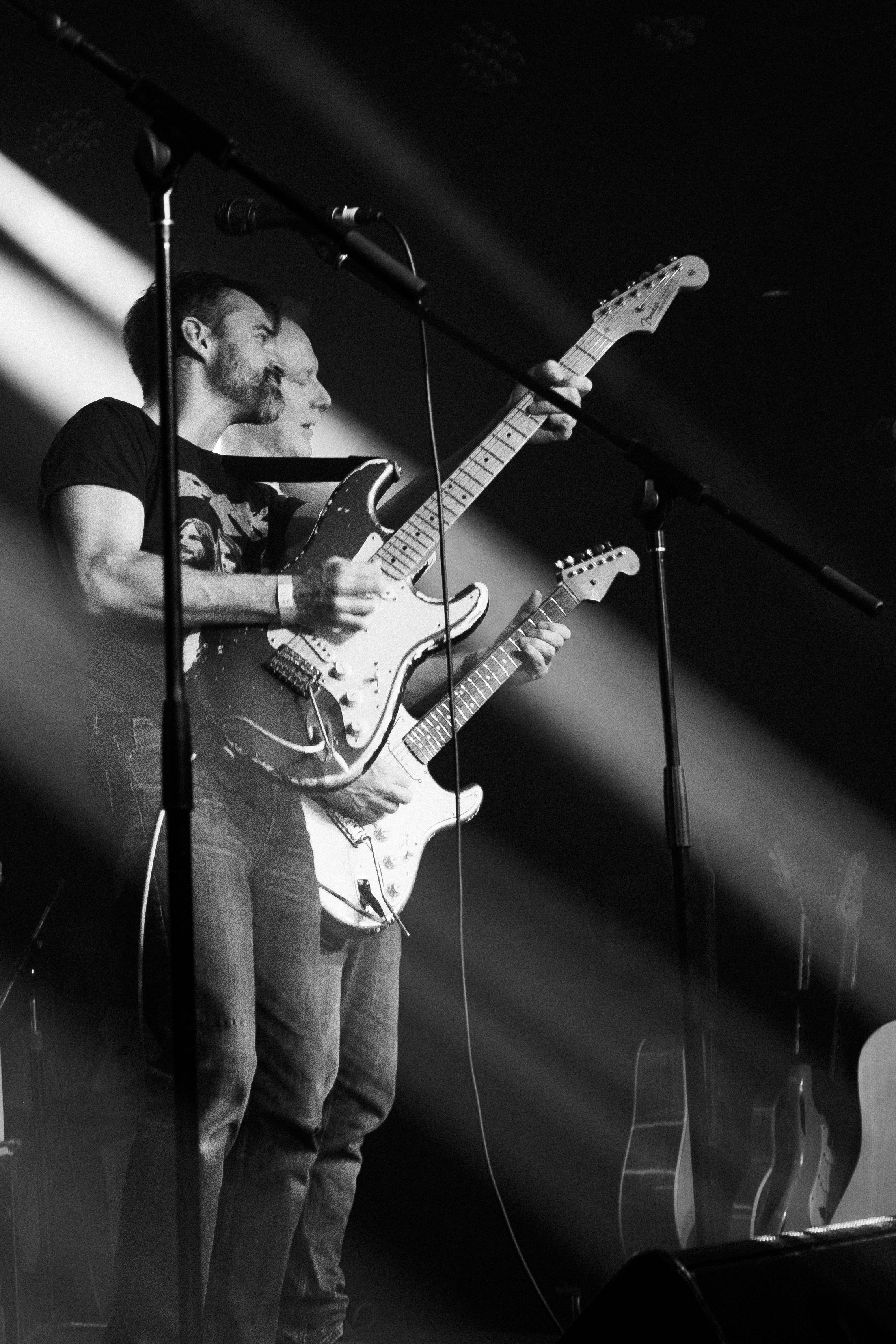 Two musicians playing electric guitars on stage during a concert, with stage lighting and microphones visible.